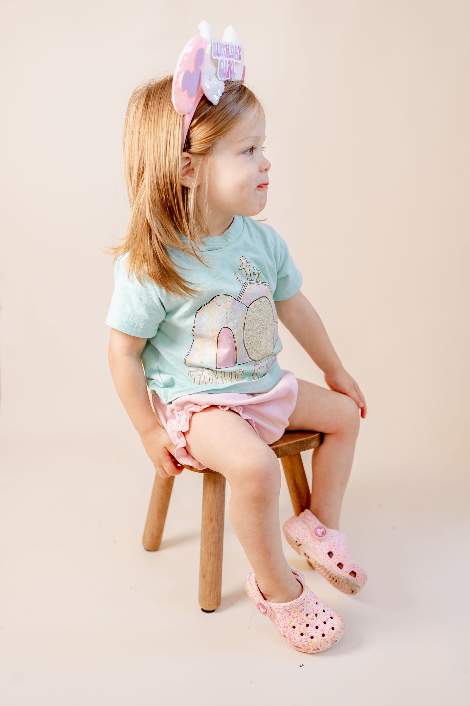 A wooden stool is positioned against a neutral background with pink and mint clothing details.