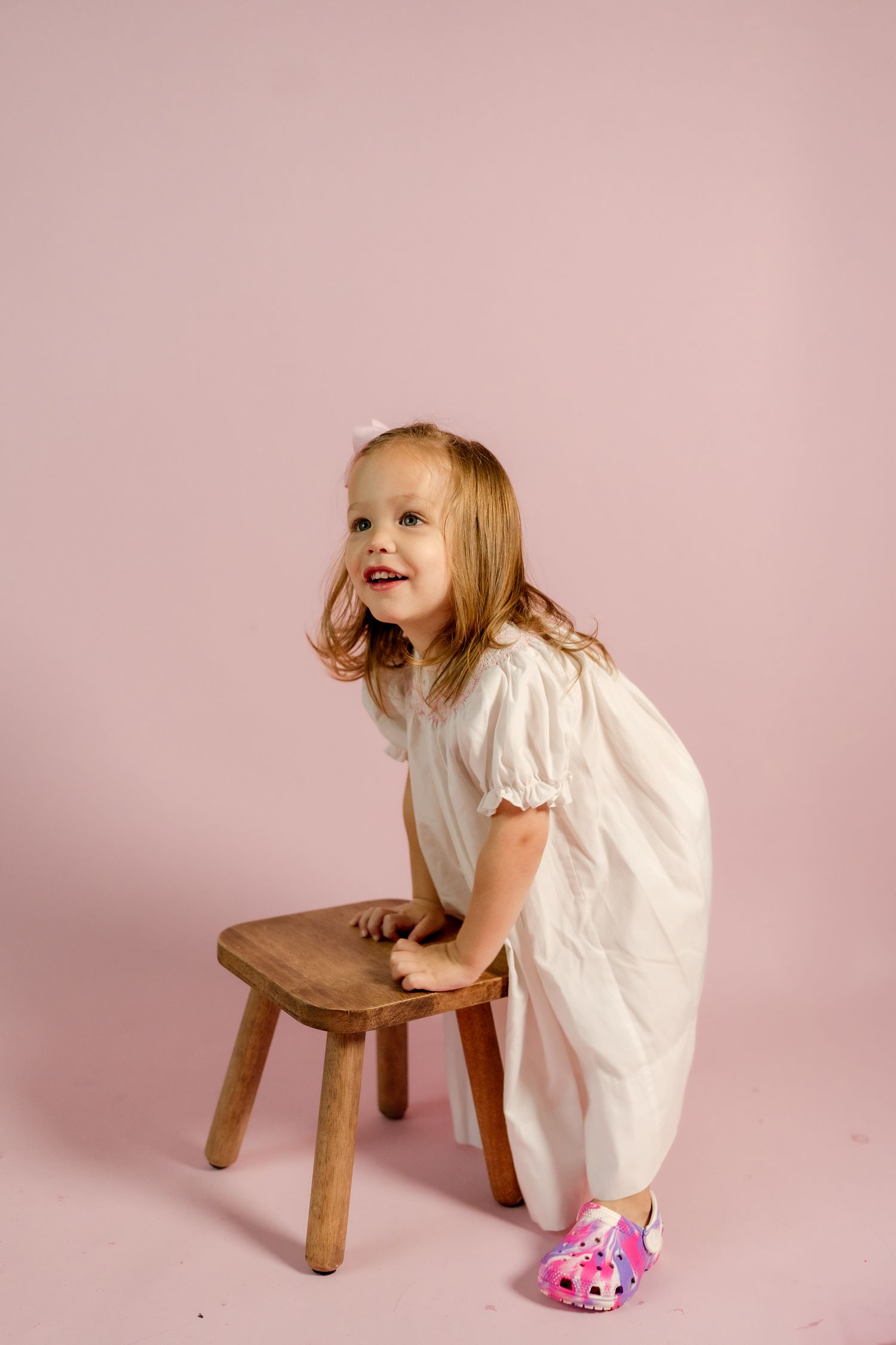 Toddler in white dress plays with wooden stool against pink backdrop in creative photoshoot sequence.