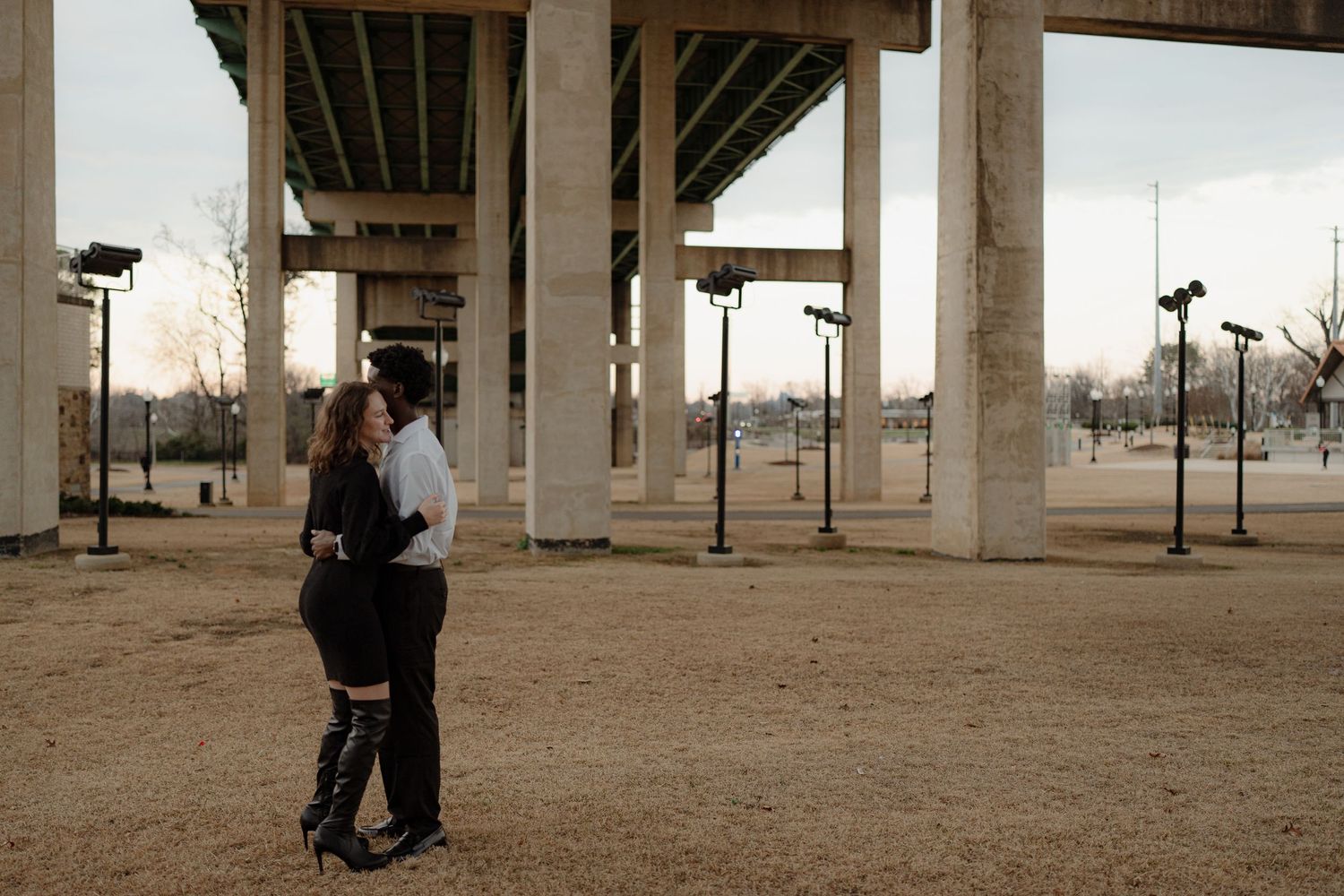 Interracial couple embracing and facing each other beneath large concrete highway overpass at dusk.