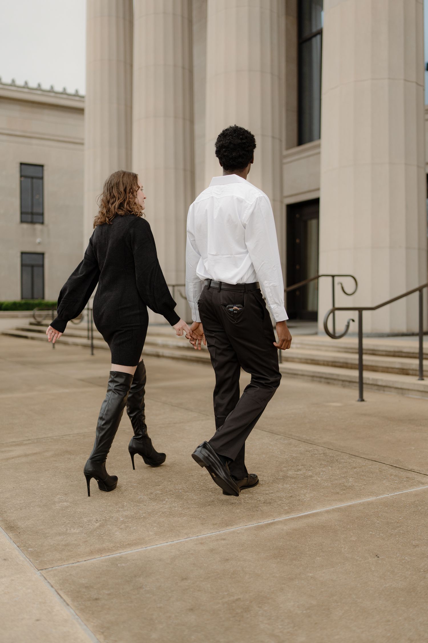 Interracial couple holding hands walking past grand columns, woman in black dress and thigh-high boots, man in white shirt.