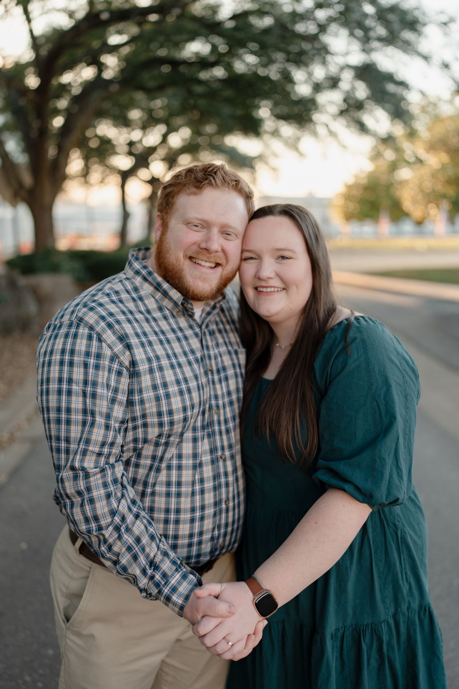 Happy couple holding hands and smiling outdoors, man in plaid shirt and woman in teal dress, golden hour park setting.
