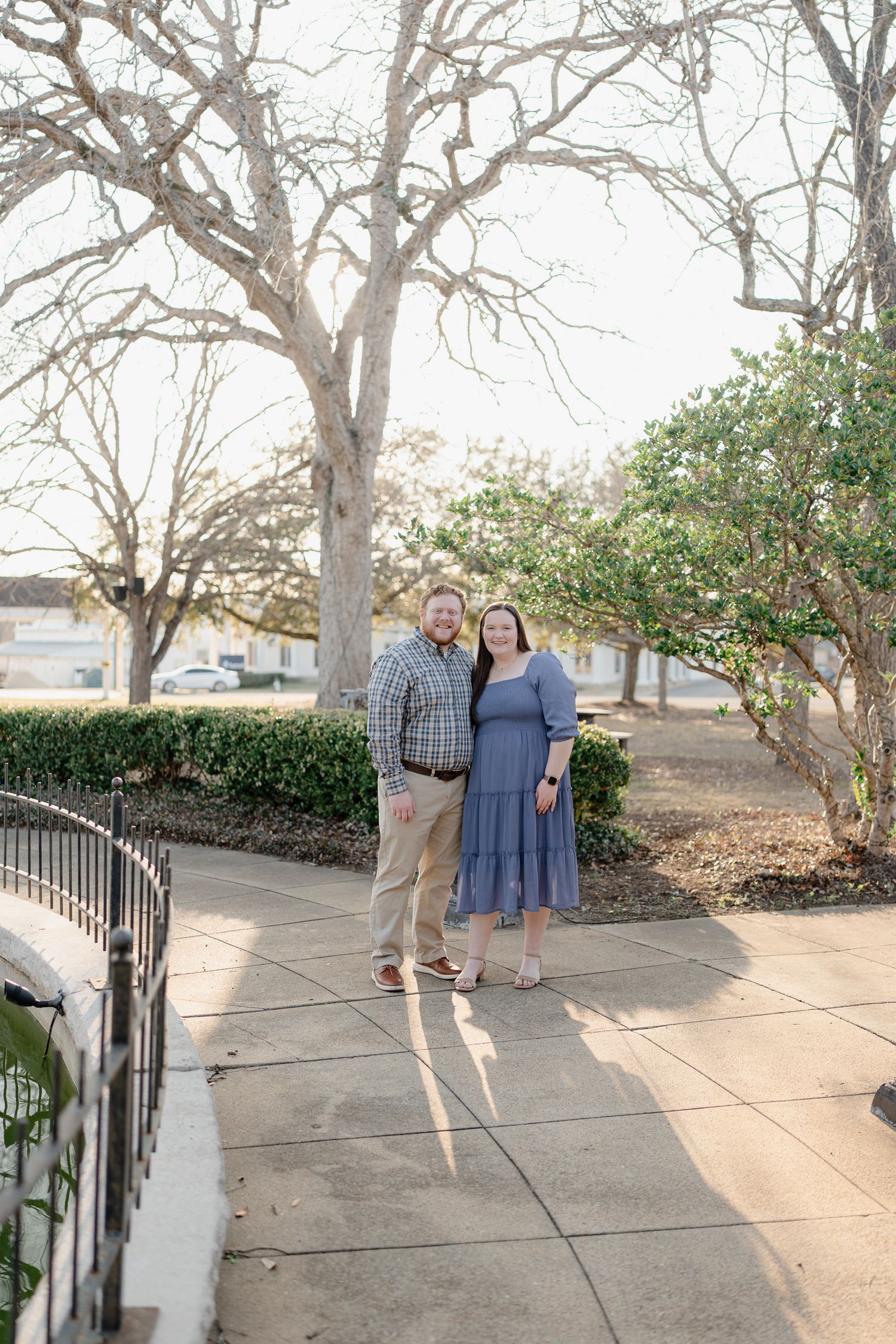 Couple posing on a sunny sidewalk surrounded by bare trees and green shrubs in an outdoor park setting.