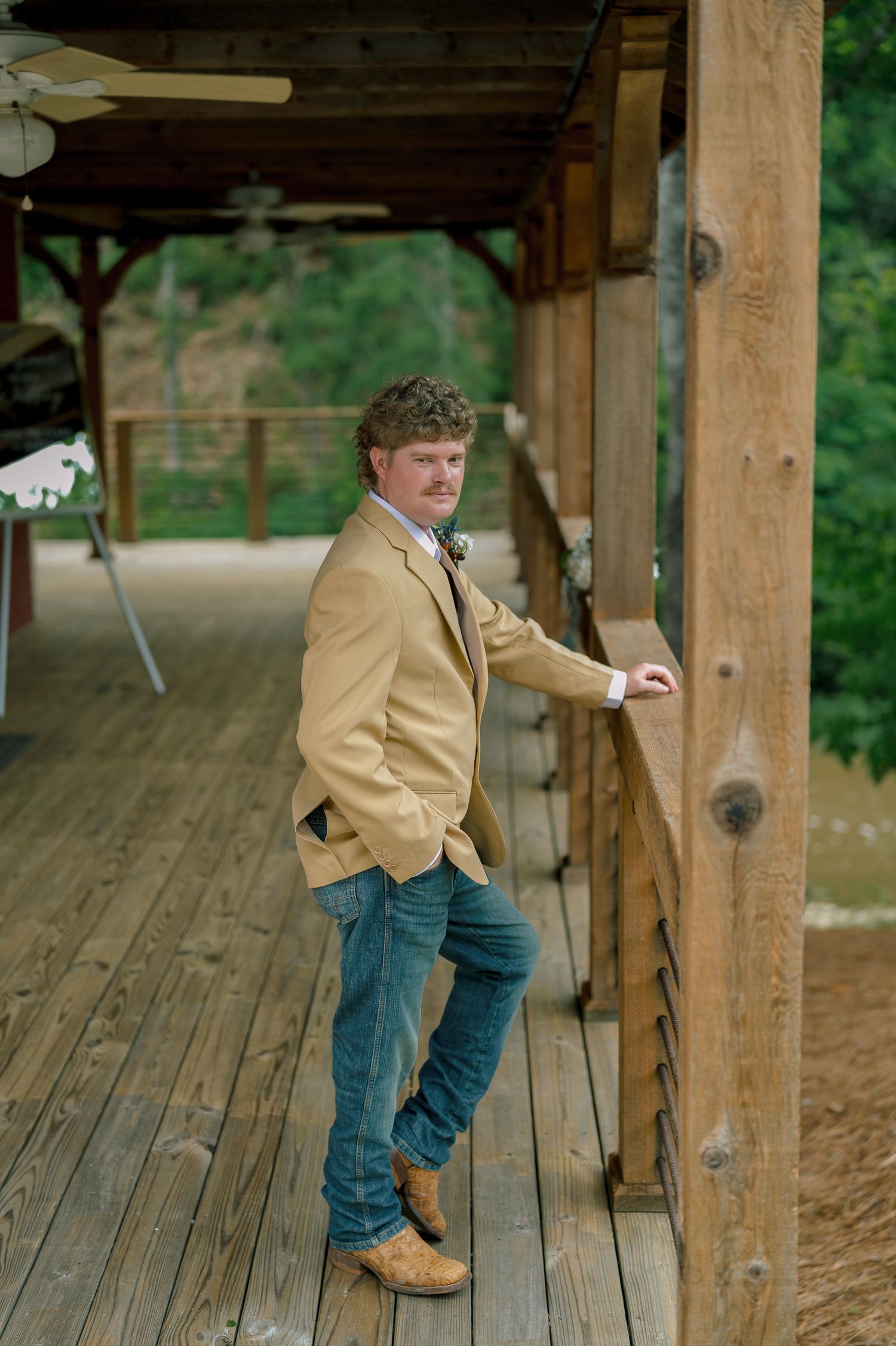 Groom in tan blazer and jeans leaning against wooden post on rustic barn venue porch.