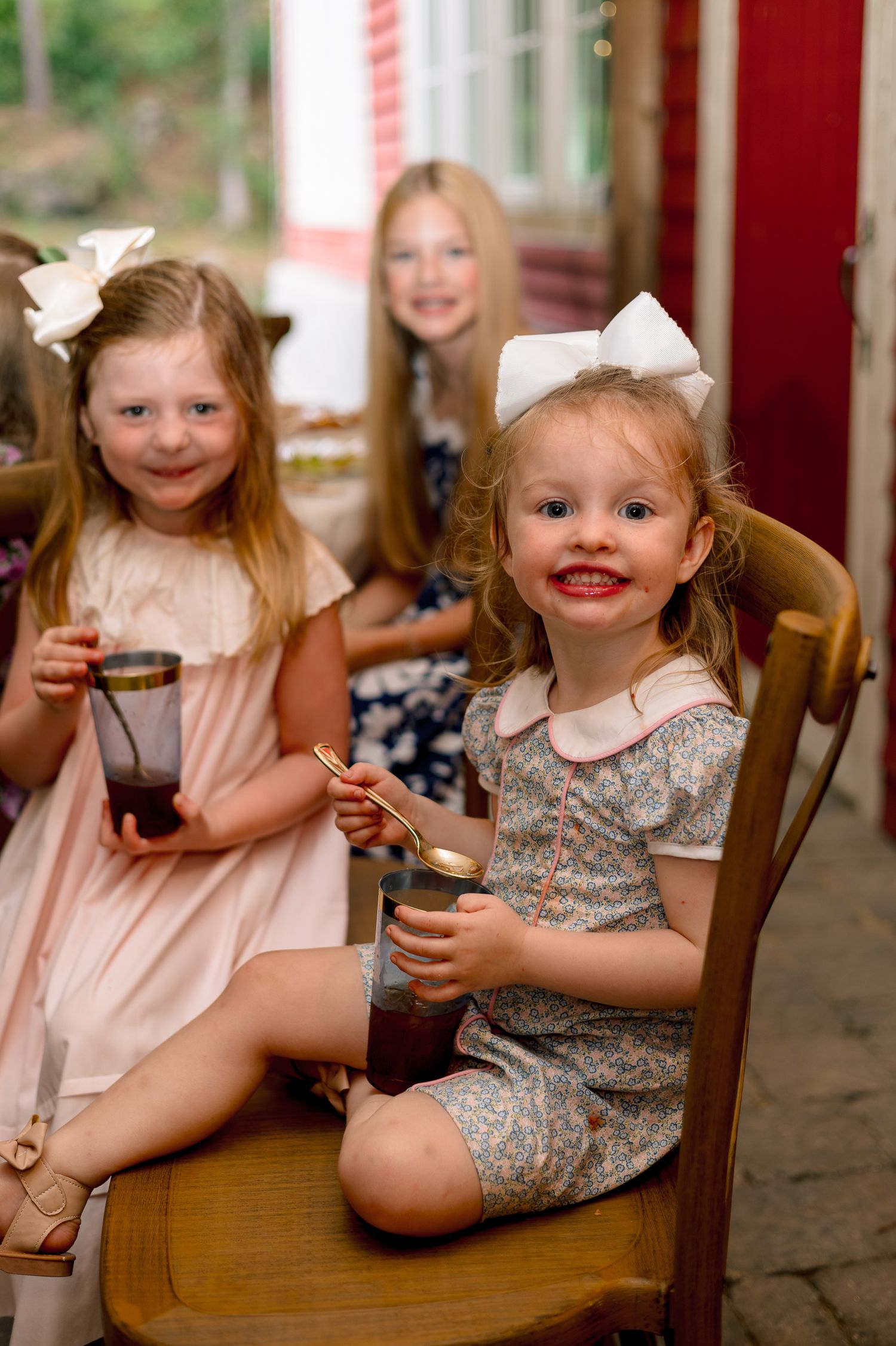 Three young girls in white hair bows enjoy dessert from mason jars at an outdoor wedding celebration.