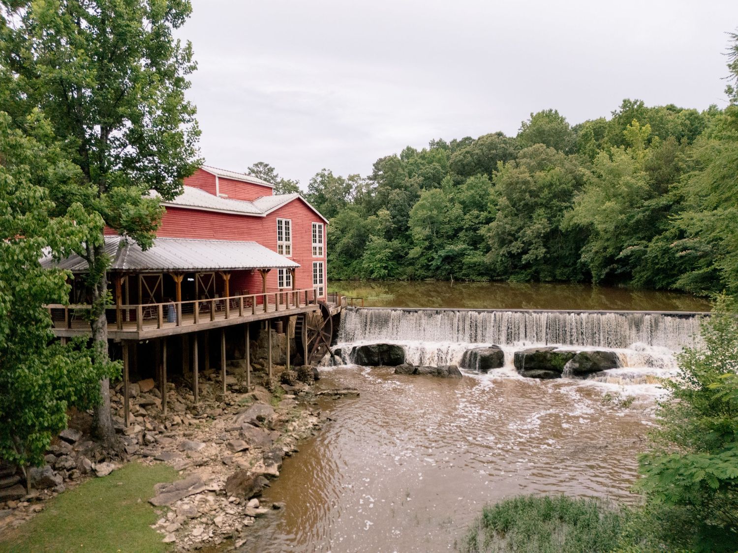 Historic red grist mill with water wheel beside cascading dam waterfall surrounded by green trees.