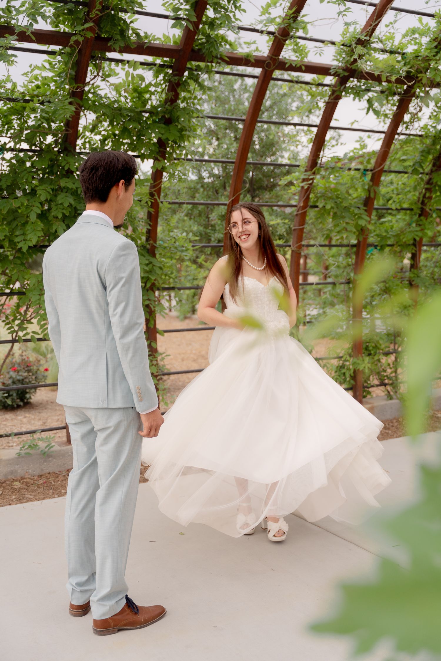 A couple in wedding attire playfully dances together in a greenhouse setting with natural light filtering through.