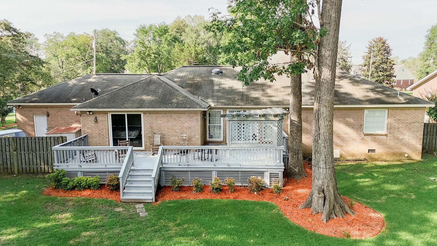 Single-story brick ranch home with wooden deck and fresh red mulch landscaping around mature trees in backyard.