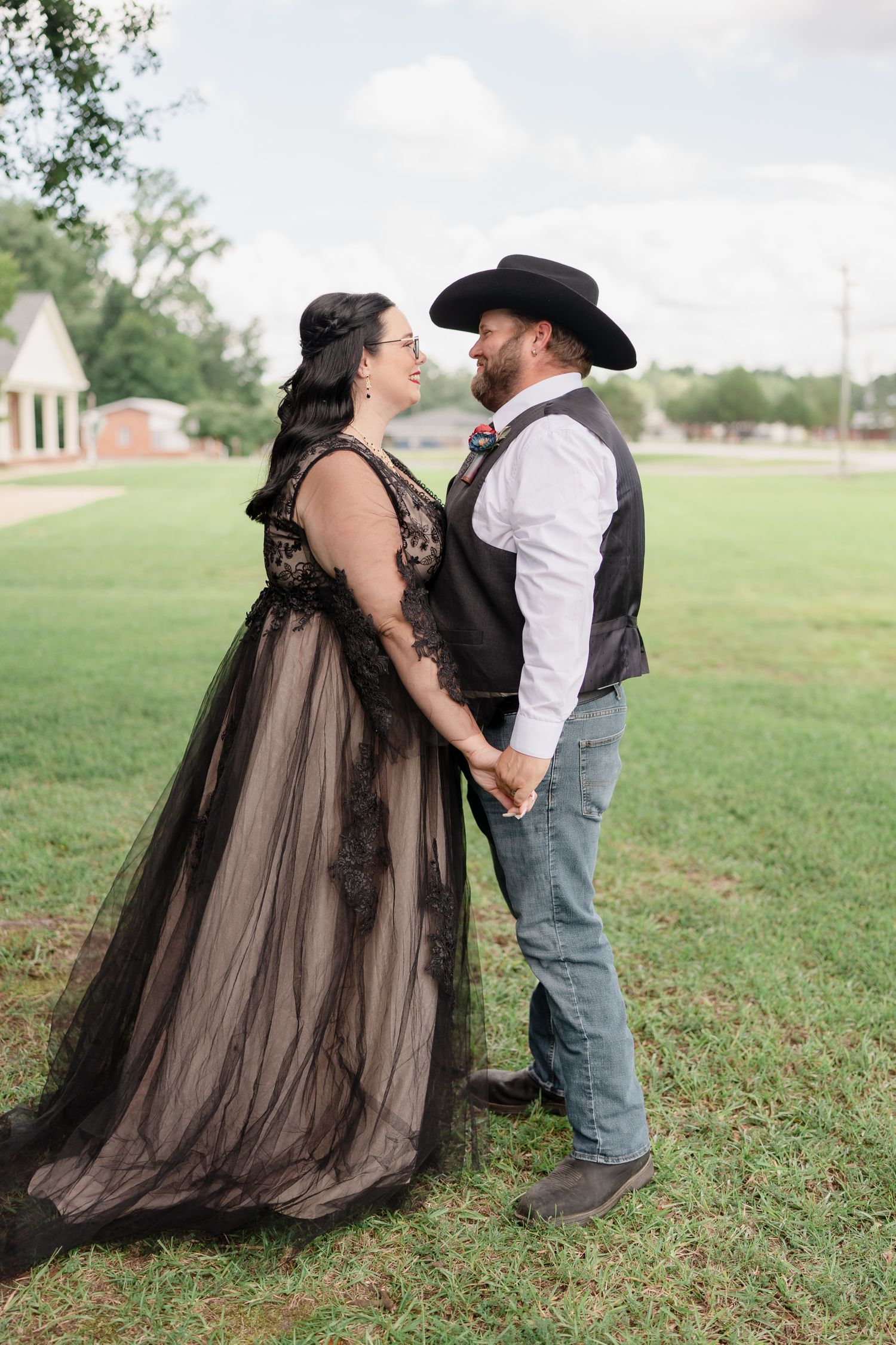 A romantic couple shares playful moments in a field wearing a black gown and cowboy attire.