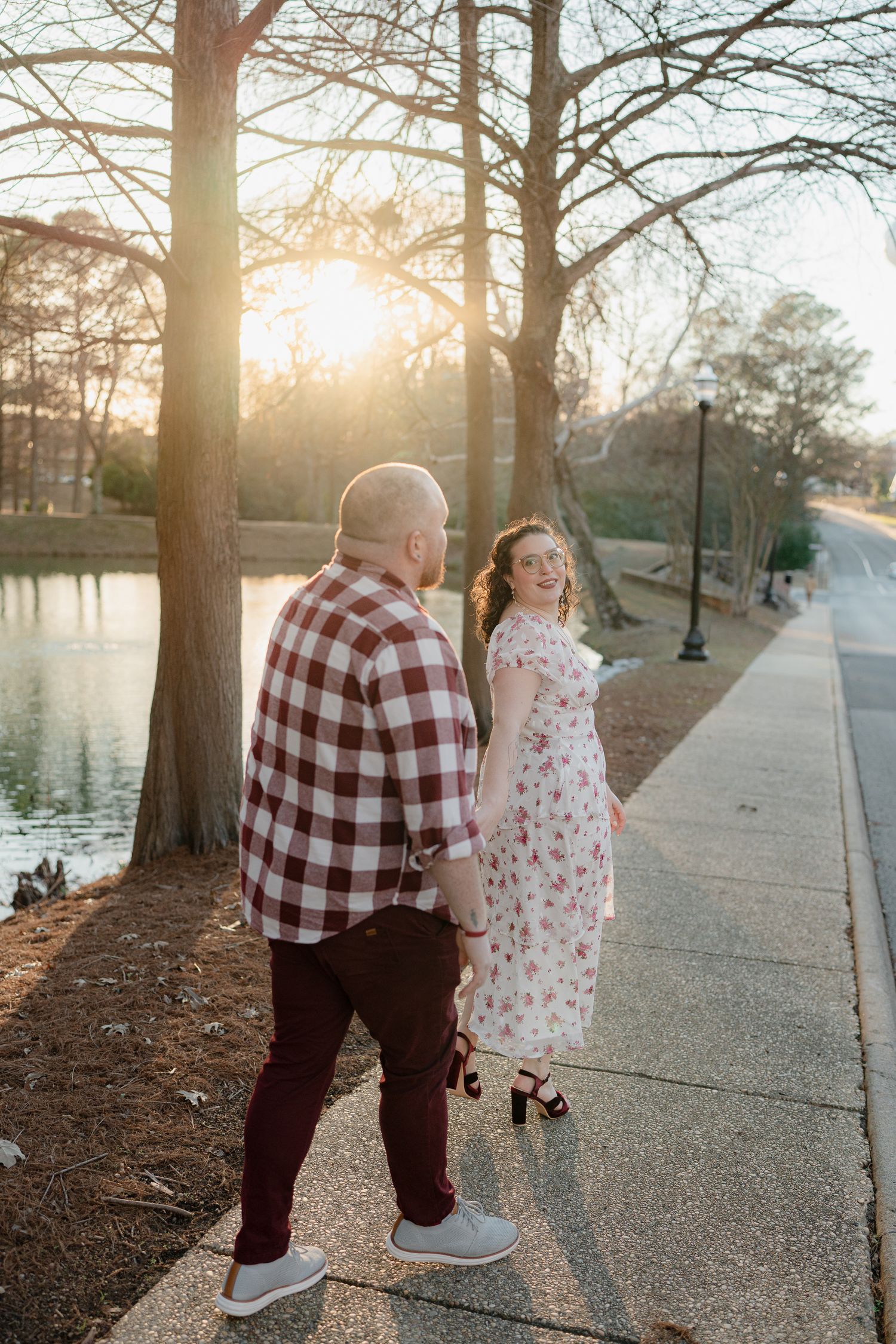 A couple poses by a lake at sunset, with sunlight filtering through trees behind them.