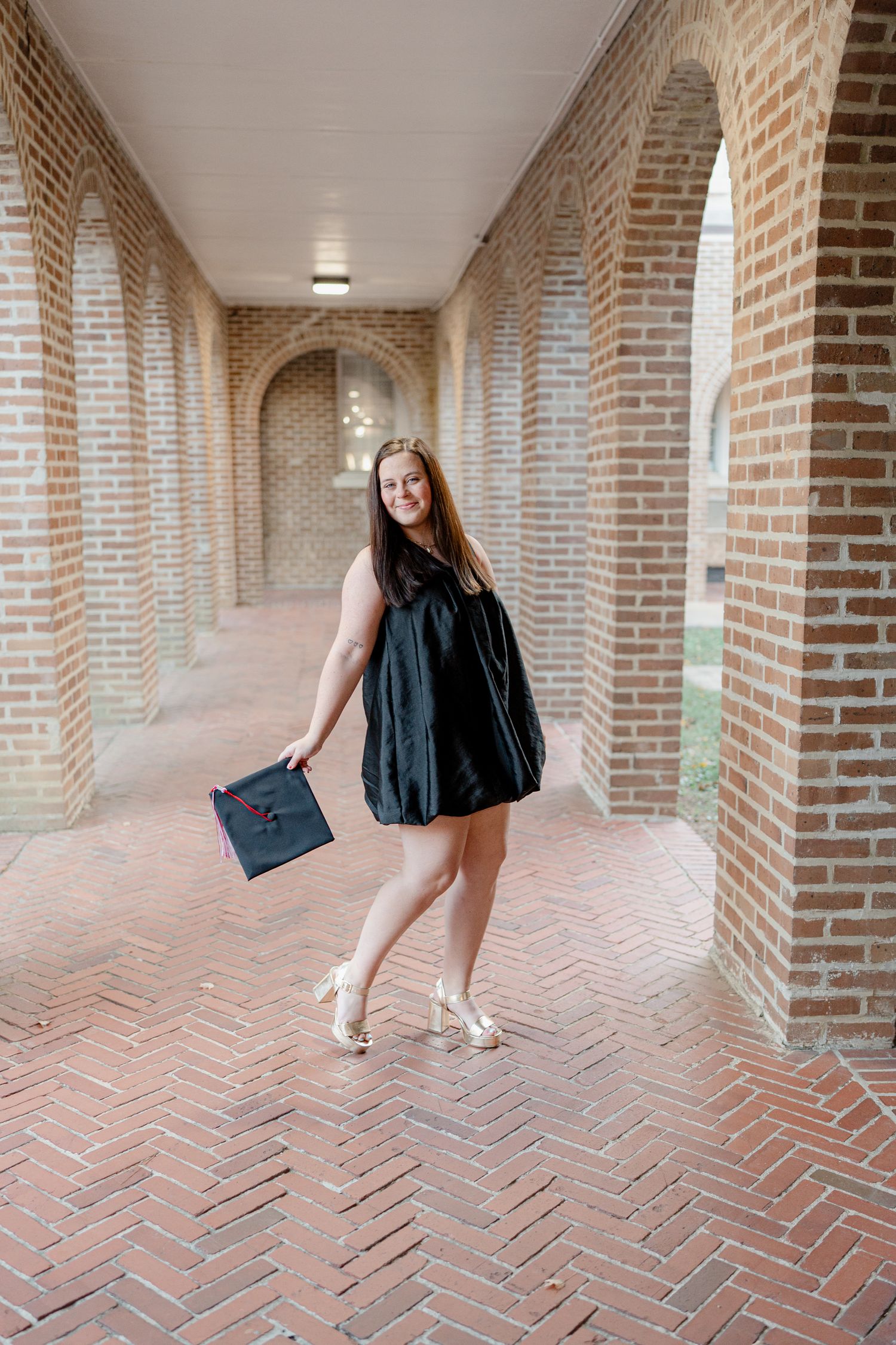 A graduate in a black dress poses in a brick archway corridor with a graduation cap.