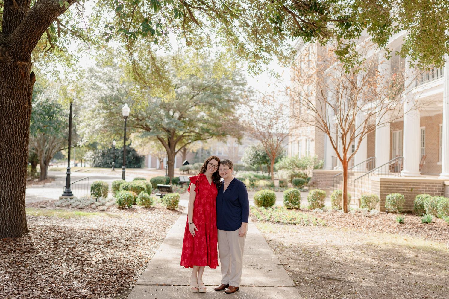 Two people in formal attire stand together on a tree-lined path outside a brick building on a sunny autumn day.