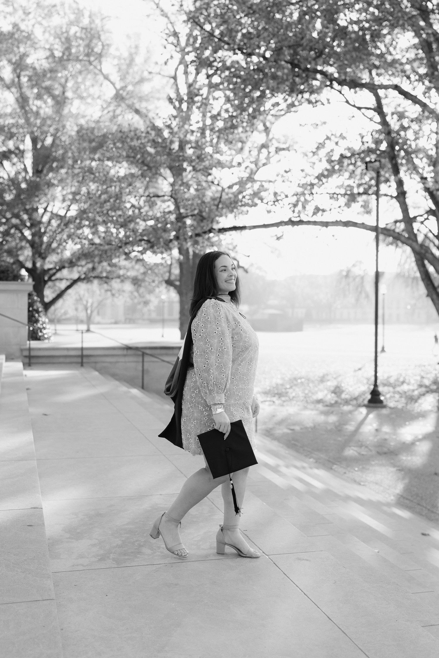 A graduate in white dress holds diploma during black and white photo session on campus.