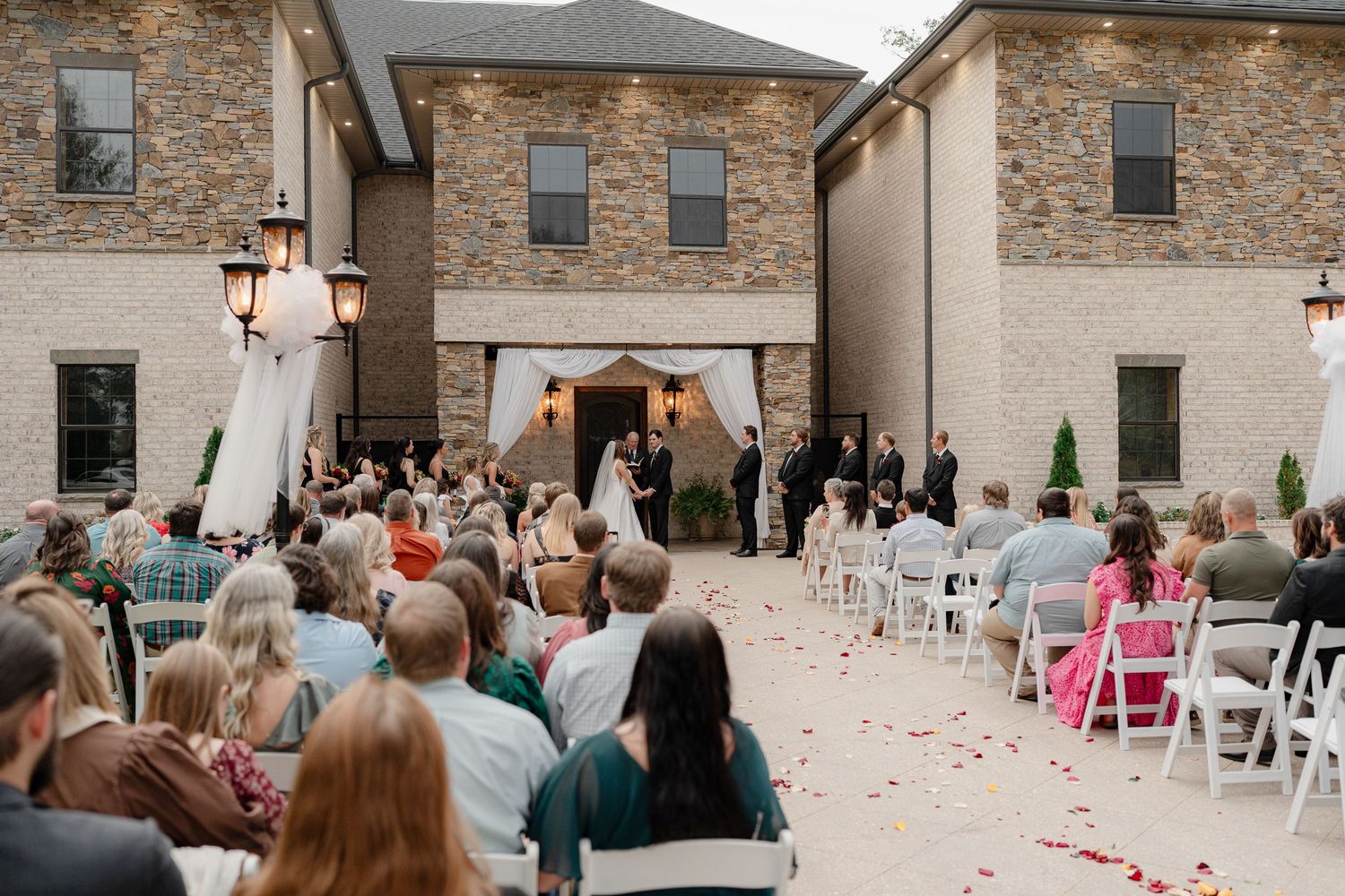 Wedding guests gather in an outdoor courtyard with stone walls and white chairs for an evening celebration.