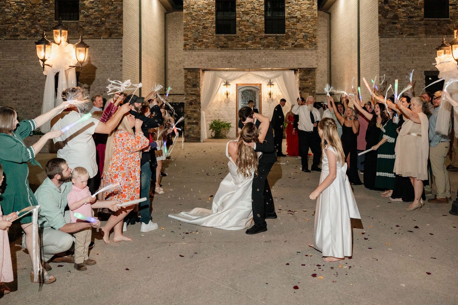 Wedding guests toss rose petals at a couple during their exit from a stone-walled venue at night.