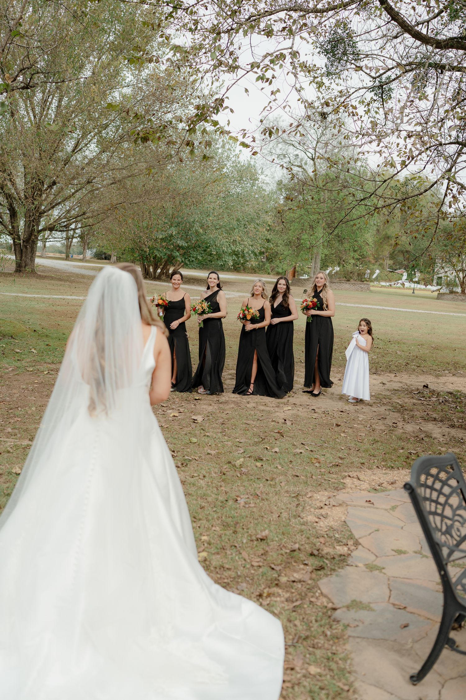 Wedding party in black dresses poses with bride in white gown and veil in outdoor autumn setting.