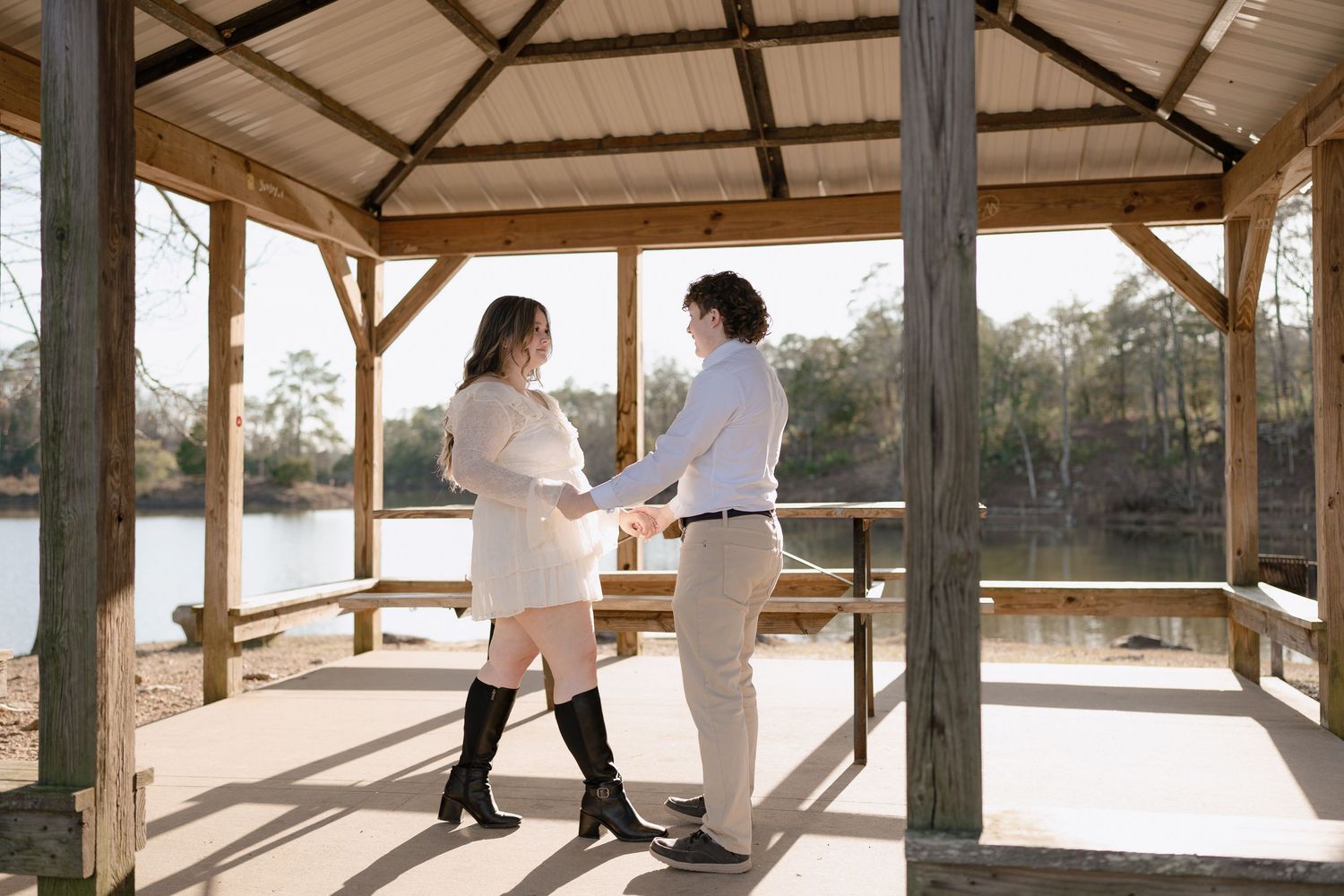 Couple holding hands in a wooden gazebo by a lake on a sunny day.