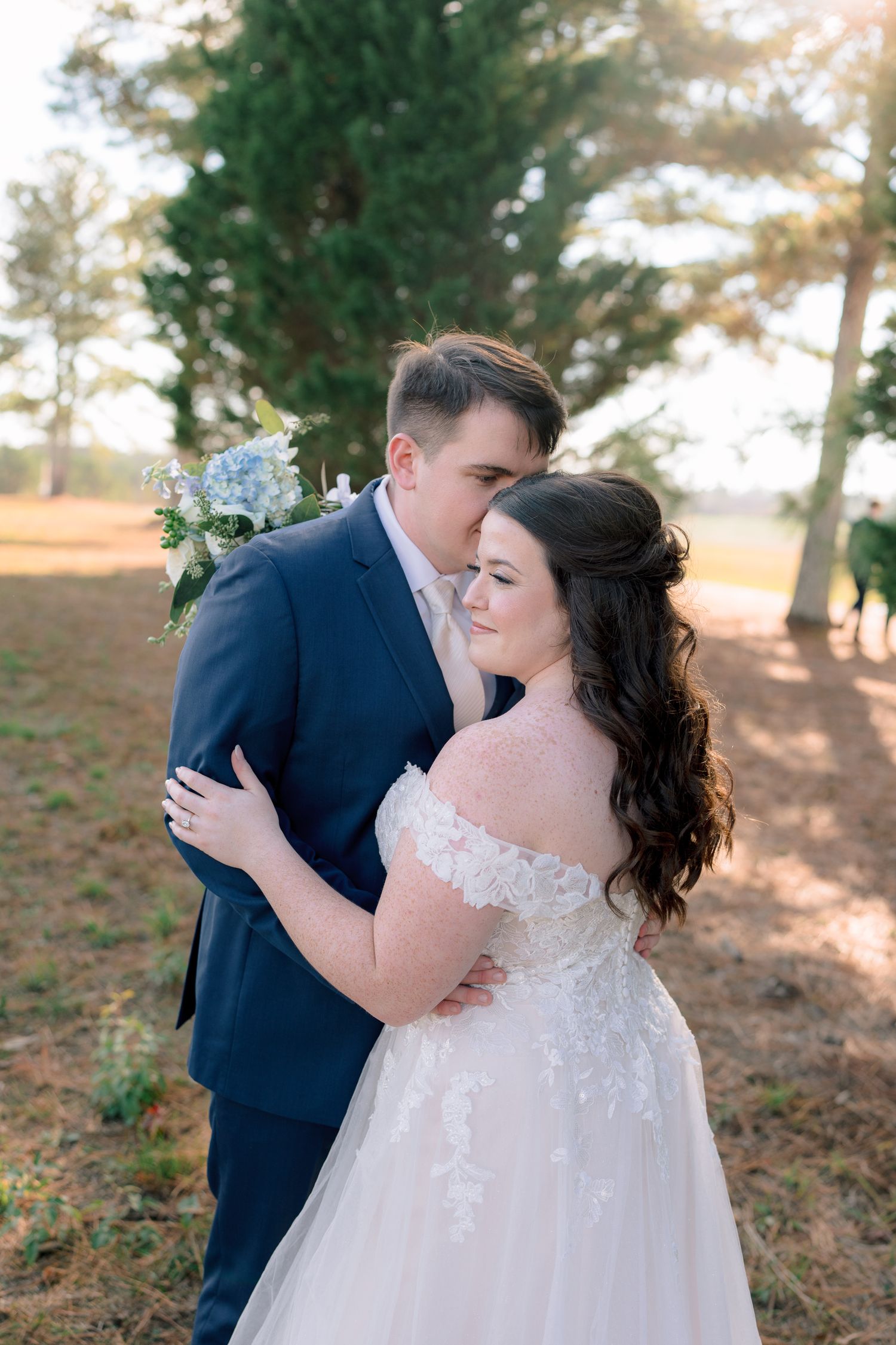 Bride in lace off-shoulder gown embraces groom in navy suit outdoors, holding blue hydrangea bouquet at golden hour.