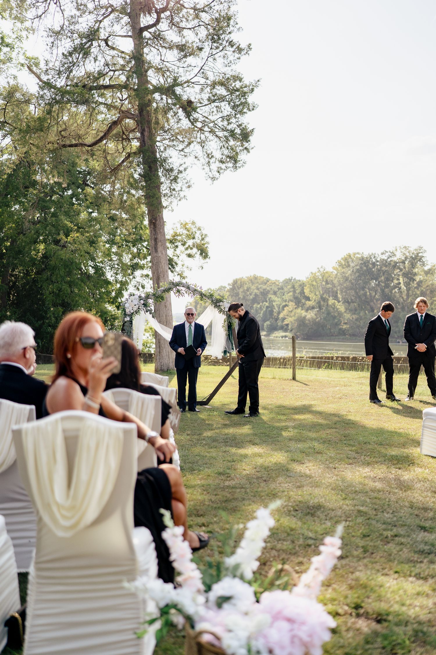 Outdoor wedding ceremony setup with white chairs and floral arrangements under tall trees on a grassy lawn.