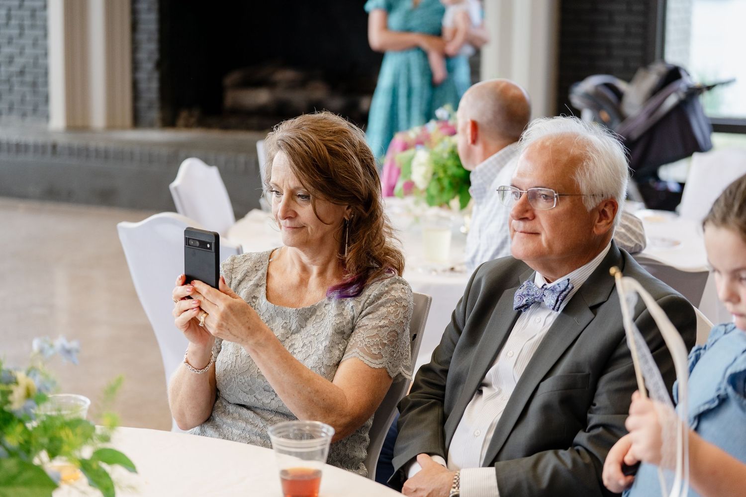 Wedding guests seated at reception tables engage in conversation and take photos during an elegant indoor celebration.