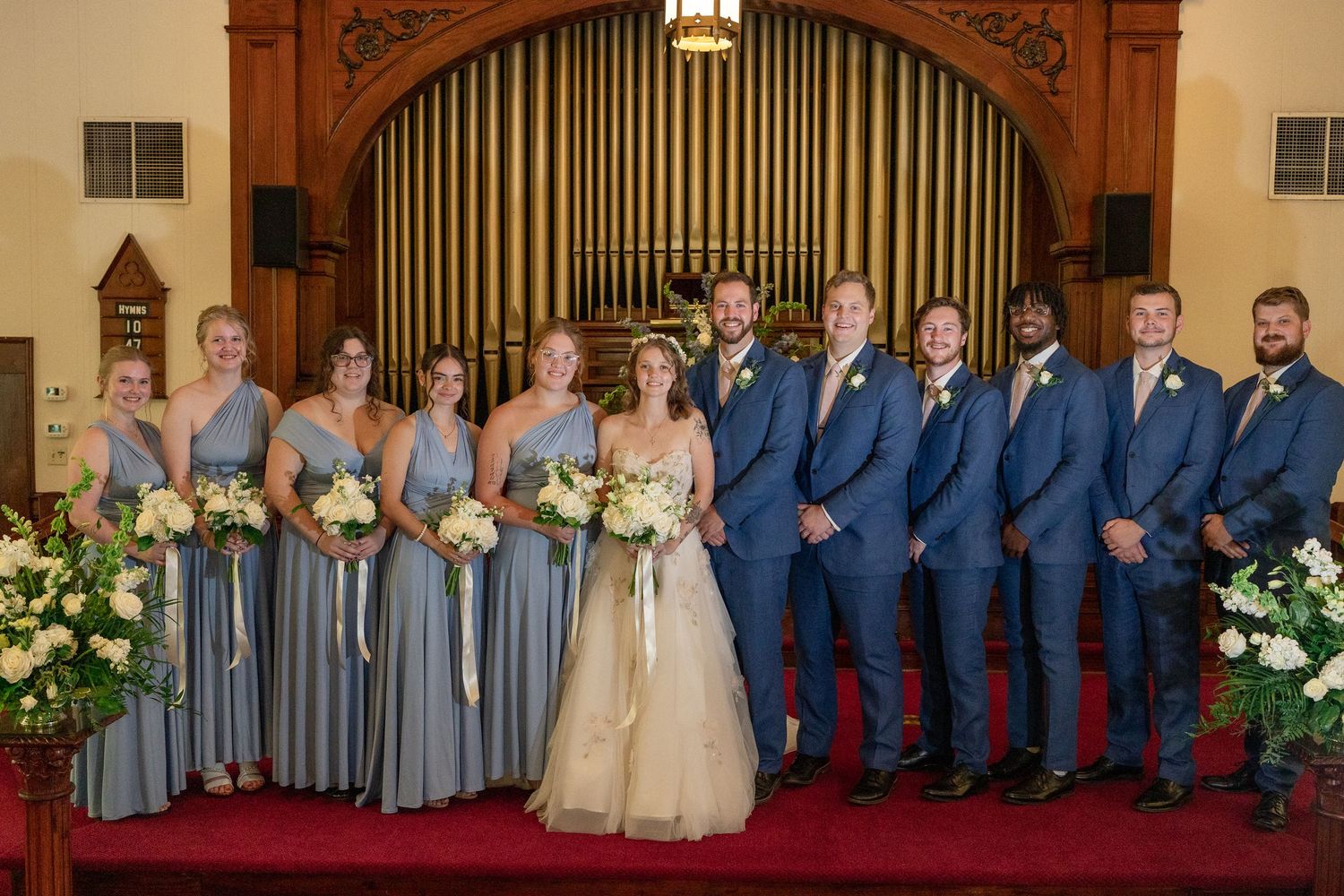 Wedding party members in blue suits and gray dresses stand together in front of ornate church organ pipes.