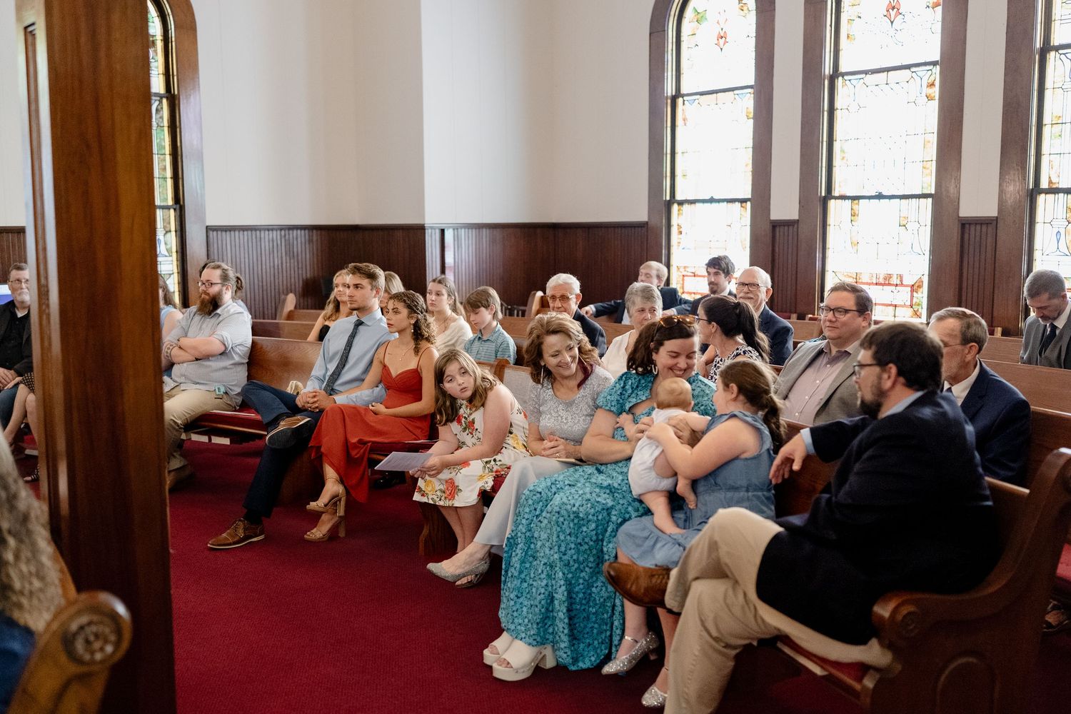Wedding guests seated in wooden pews inside a traditional church with stained glass windows and red carpeting.