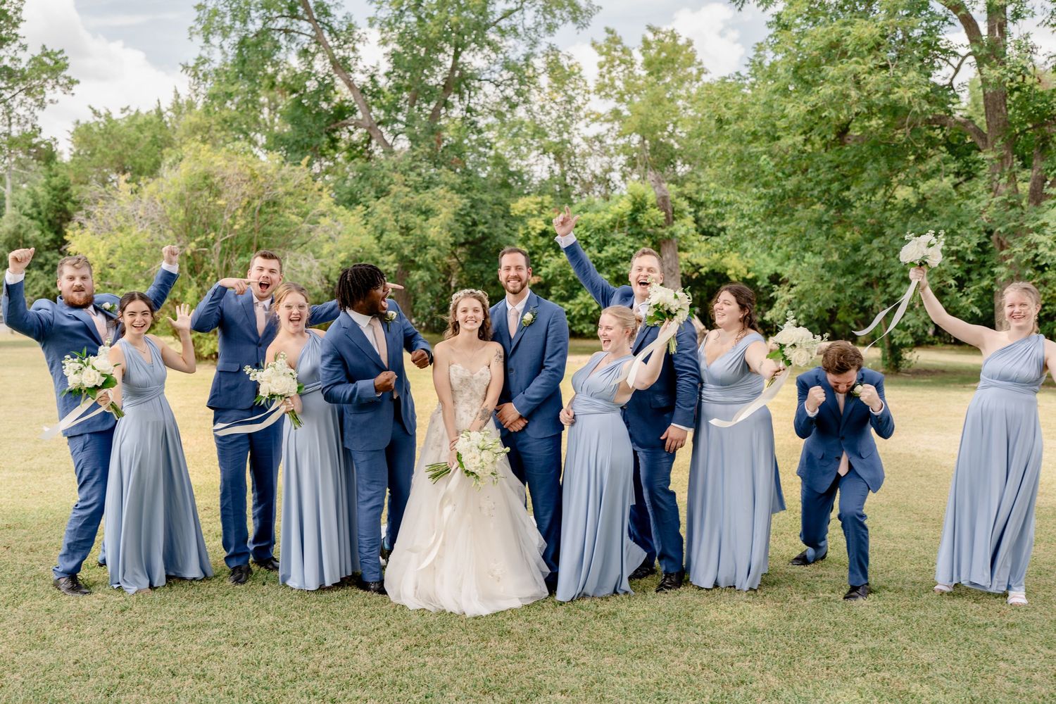 Wedding group poses casually on grass wearing coordinated blue dresses and suits in natural setting.