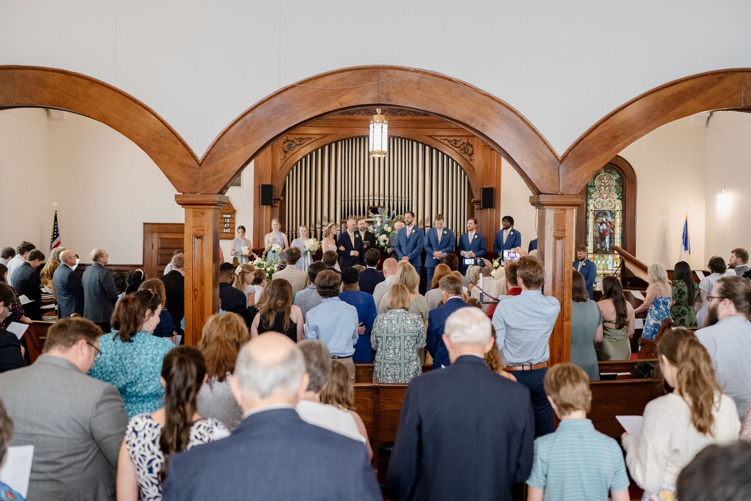 Wedding ceremony guests stand in a church with arched wooden beams and organ pipes visible in the background.