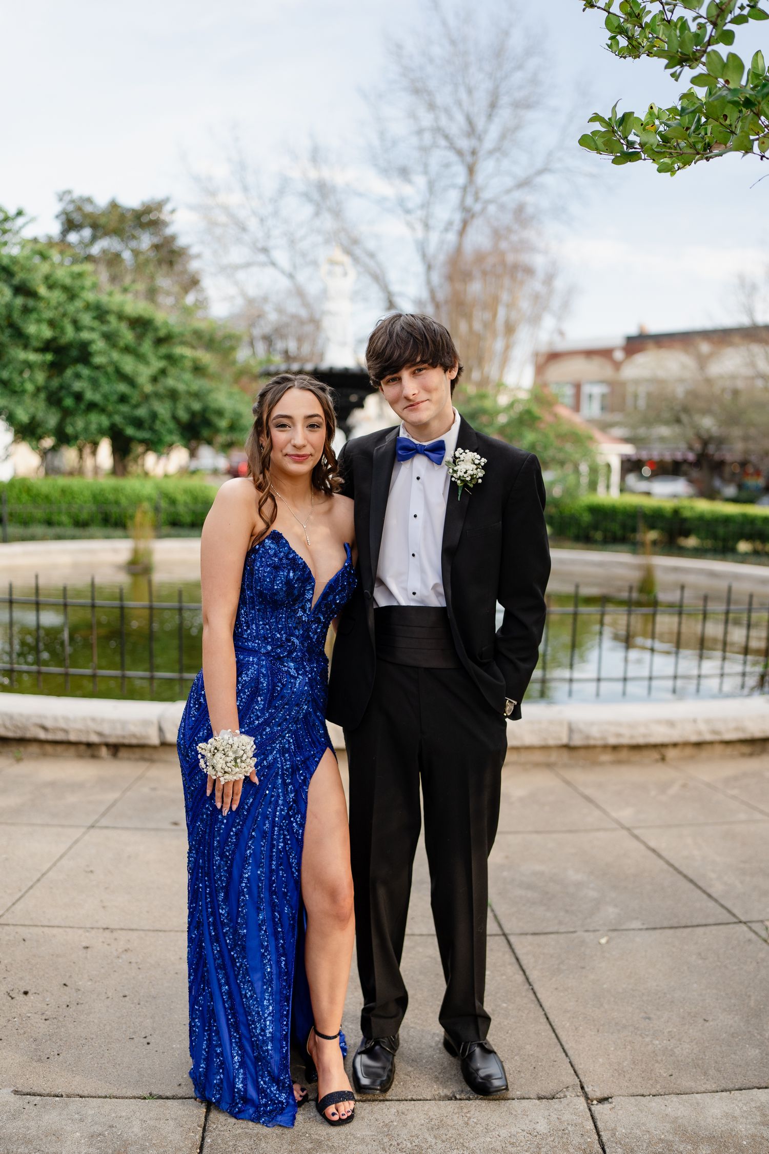 Couple in formal attire poses for photo near garden fountain in elegant outdoor setting.