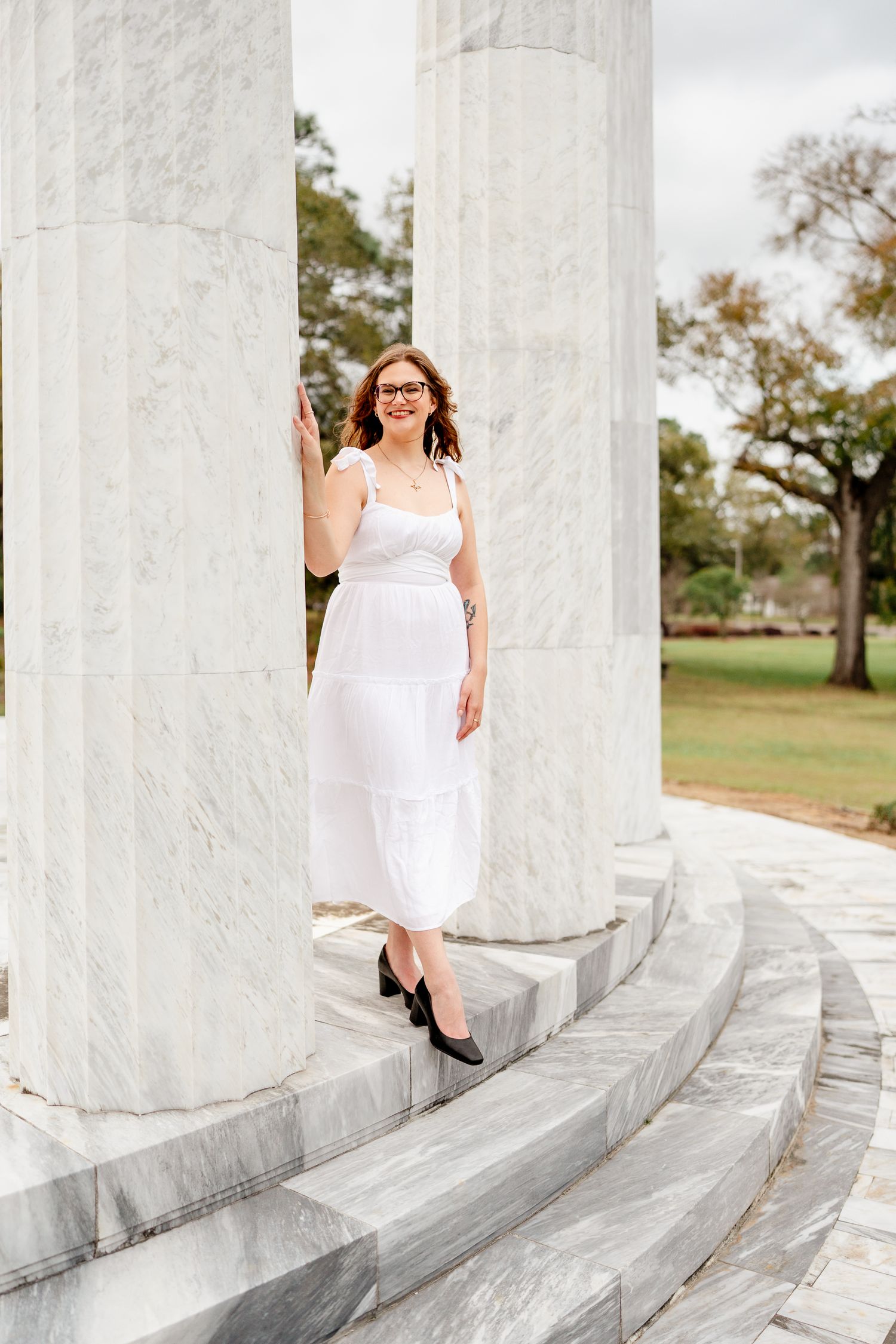 Graduate posing on marble steps in white dress and glasses against vibrant outdoor background.