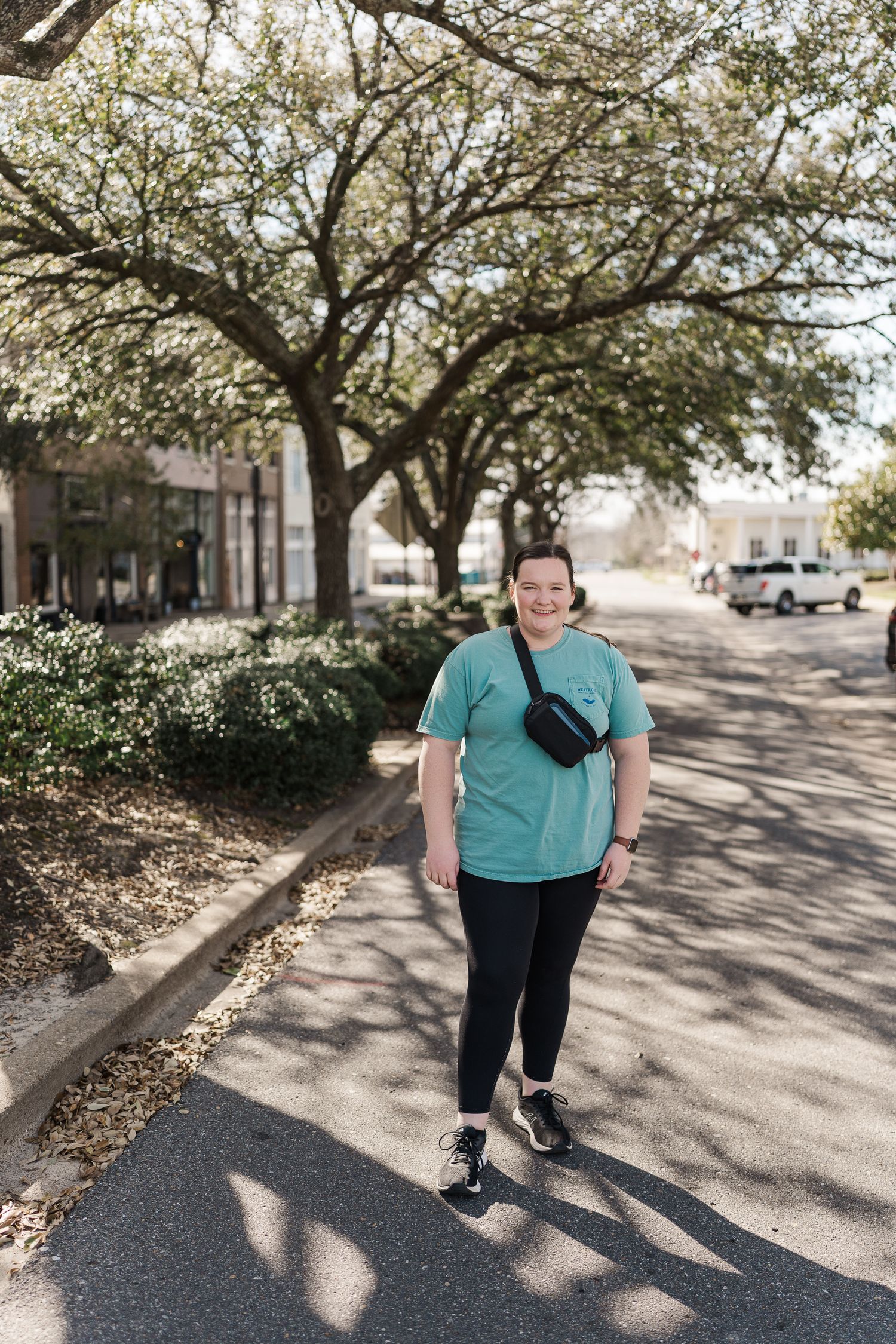 Person in teal shirt walking on tree-lined street with fanny pack.