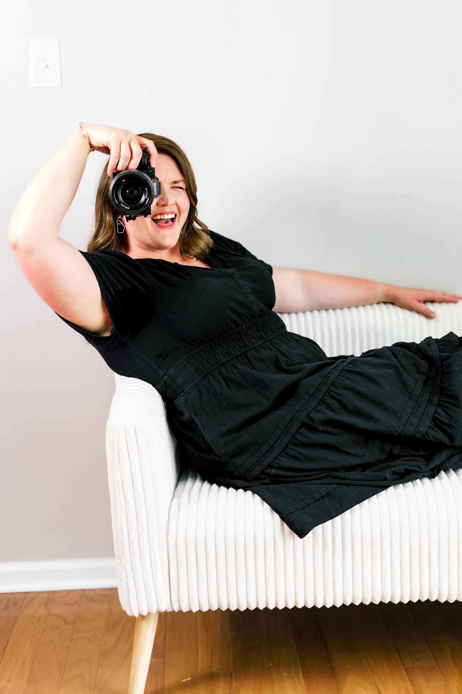 A relaxed photographer lounges on a white couch while holding a camera in a playful photo series.