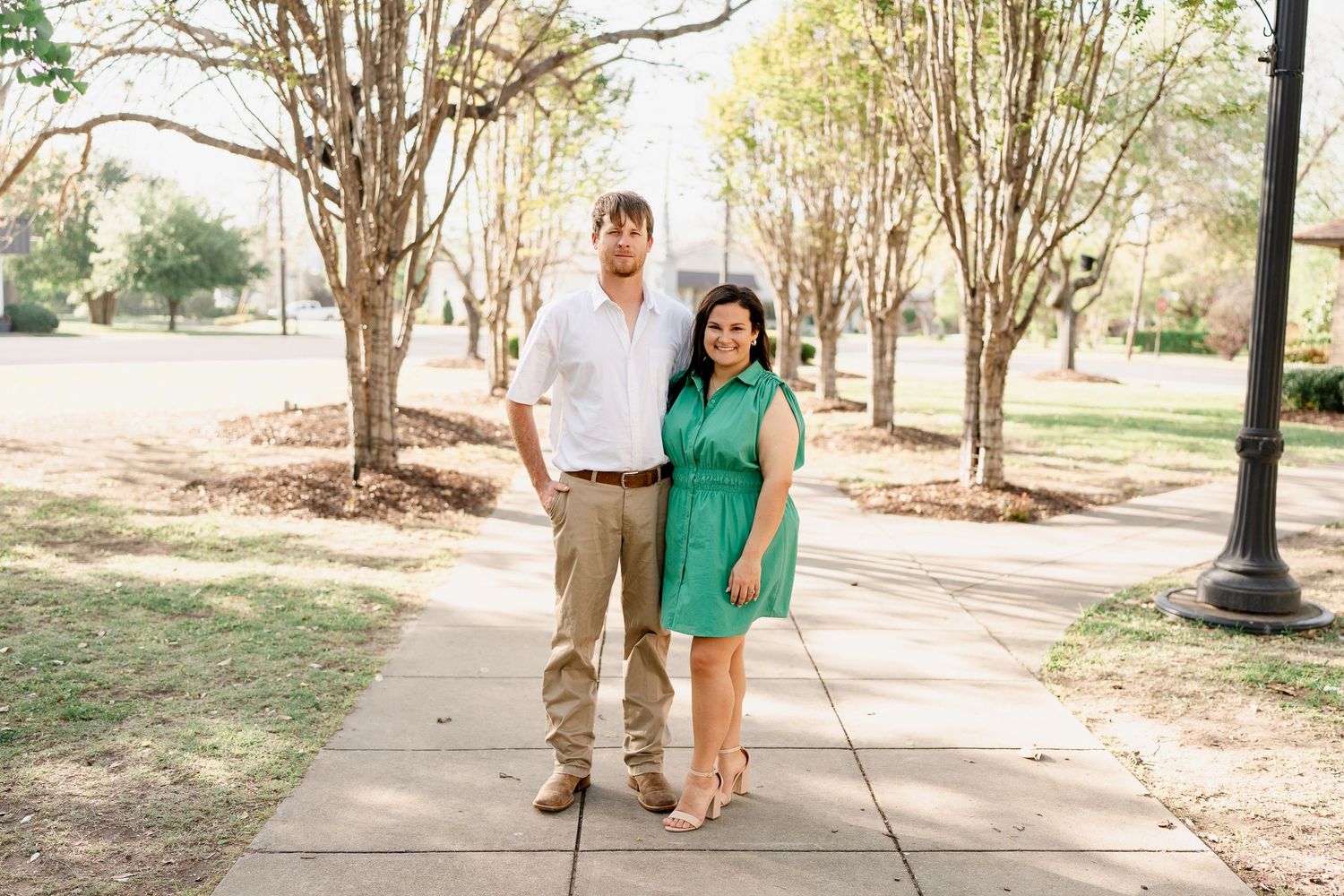 Couple stands together on a sidewalk lined with tall trees.