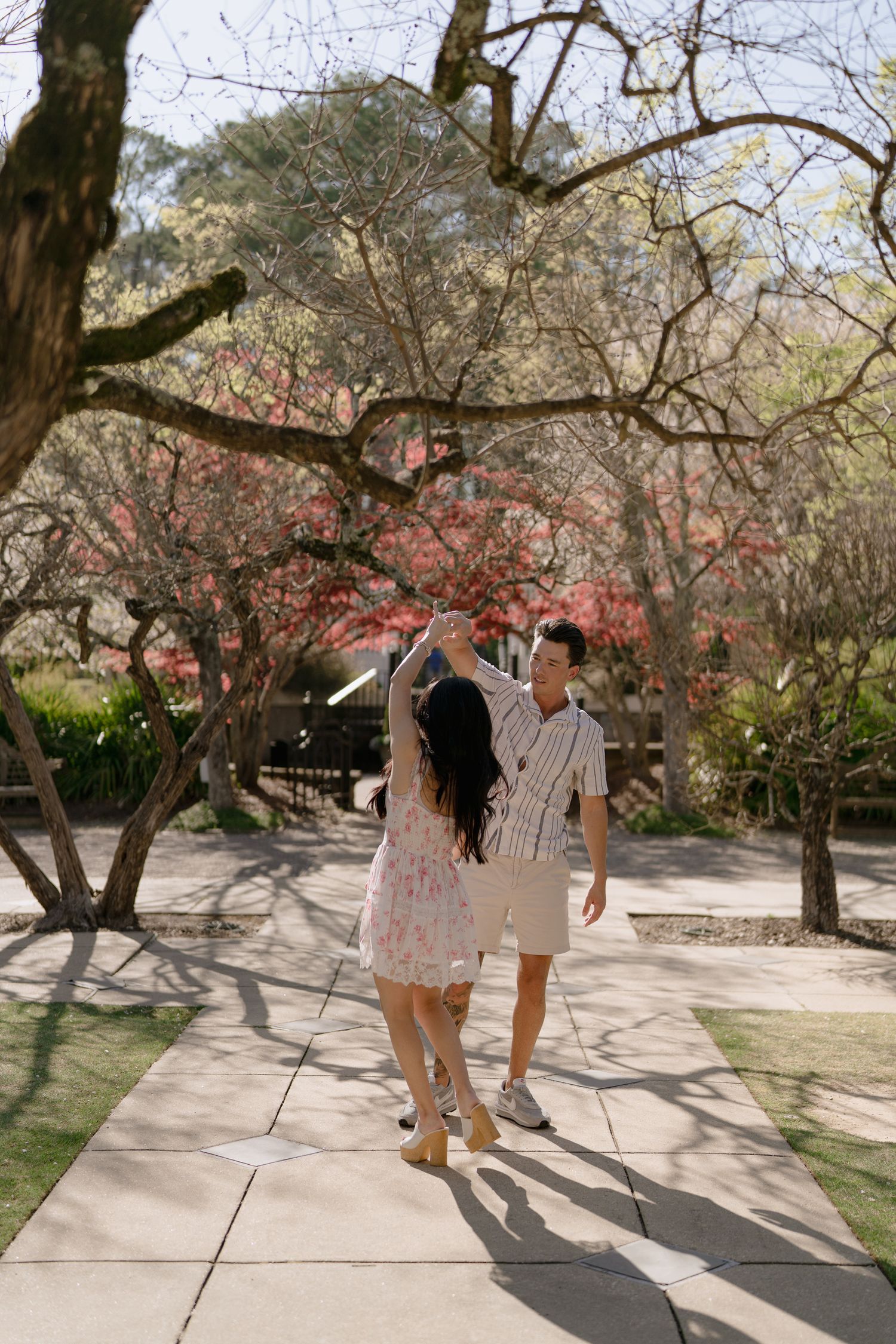 Couple dancing joyfully on a sunlit garden path with colorful blooming trees in the background.