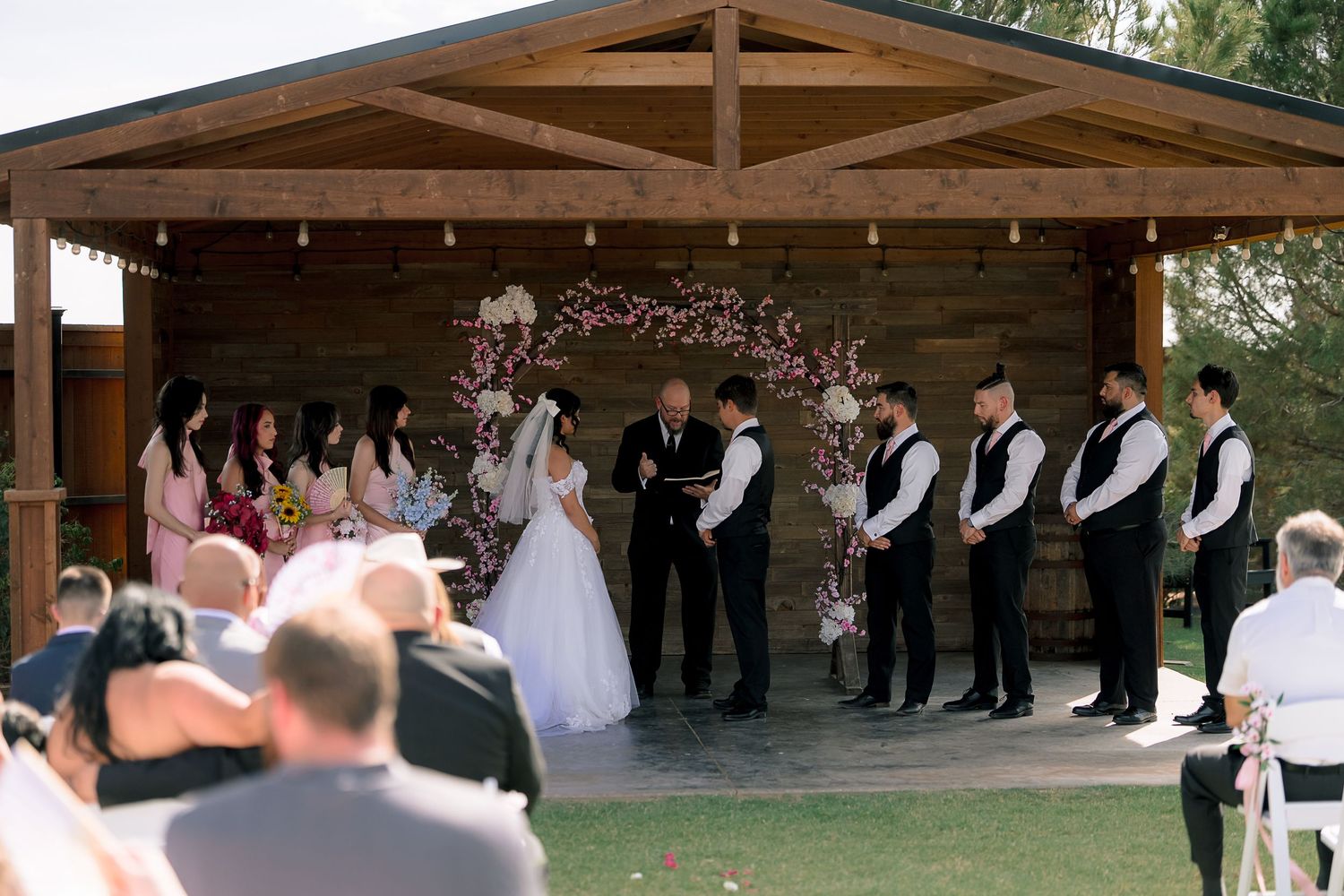 Wedding ceremony under rustic wooden pavilion with floral arch, officiant, bride, groom, and wedding party.