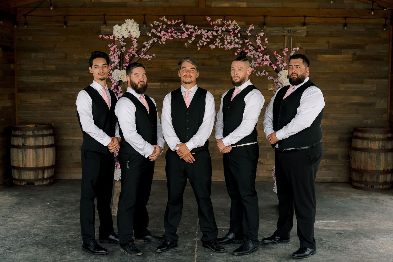 Five groomsmen in black vests and pink ties pose in a rustic barn with floral arch backdrop.