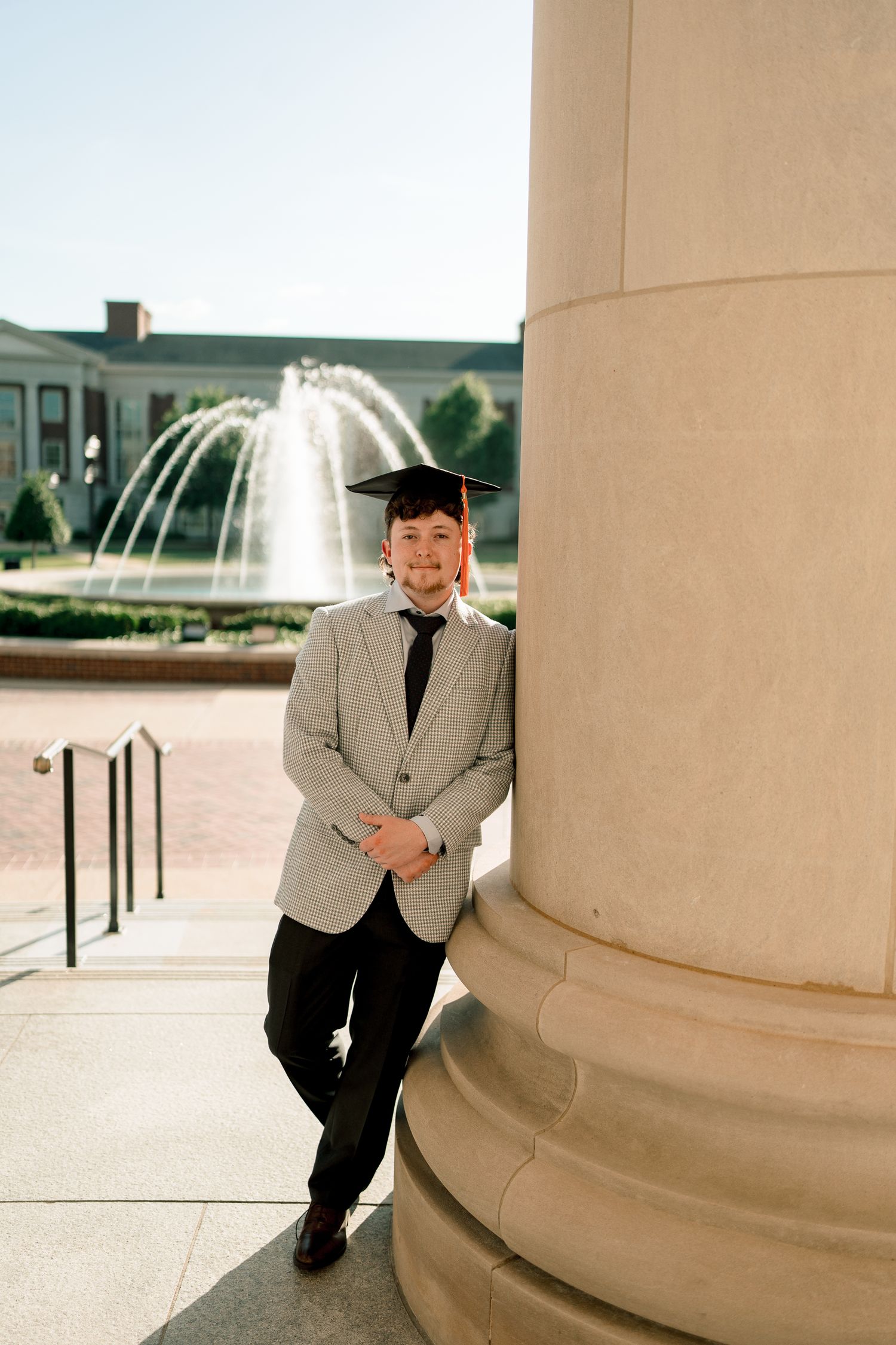 Graduate in gray blazer and mortarboard cap leans against column with fountain in background on campus.