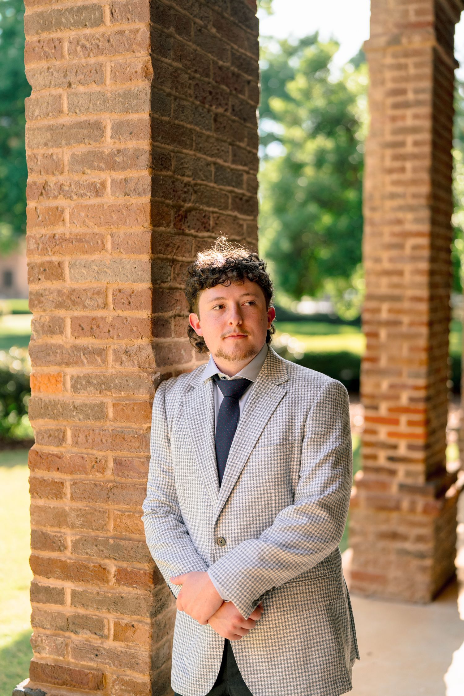 Young man in gray houndstooth blazer and tie leans against brick column on university campus walkway.