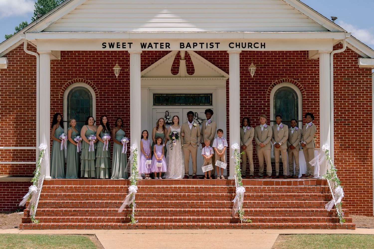 Wedding party posed on steps of Sweet Water Baptist Church, bridesmaids in sage green, groomsmen in tan suits.