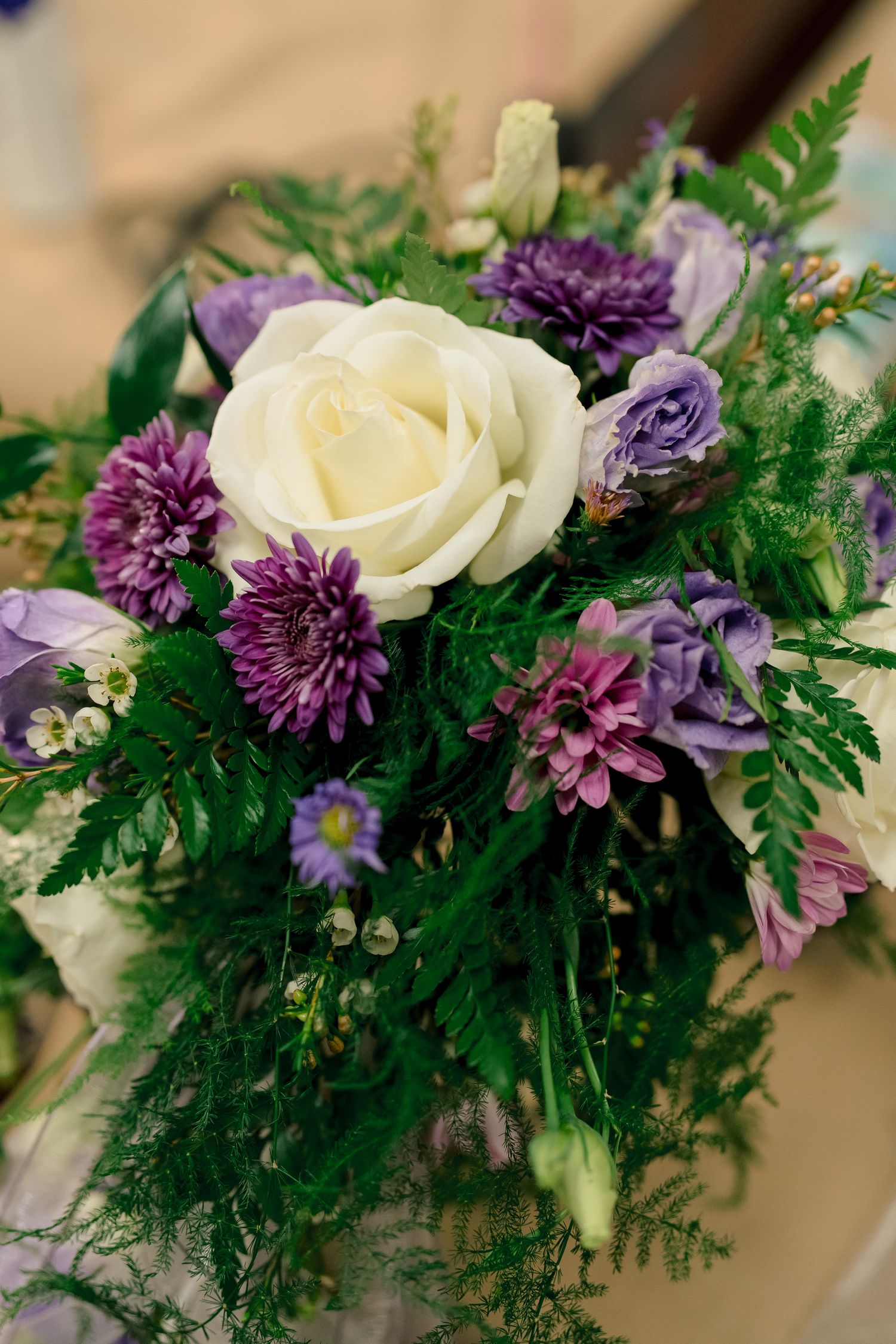 Elegant bridal bouquet featuring a white rose surrounded by purple chrysanthemums and lush green ferns.