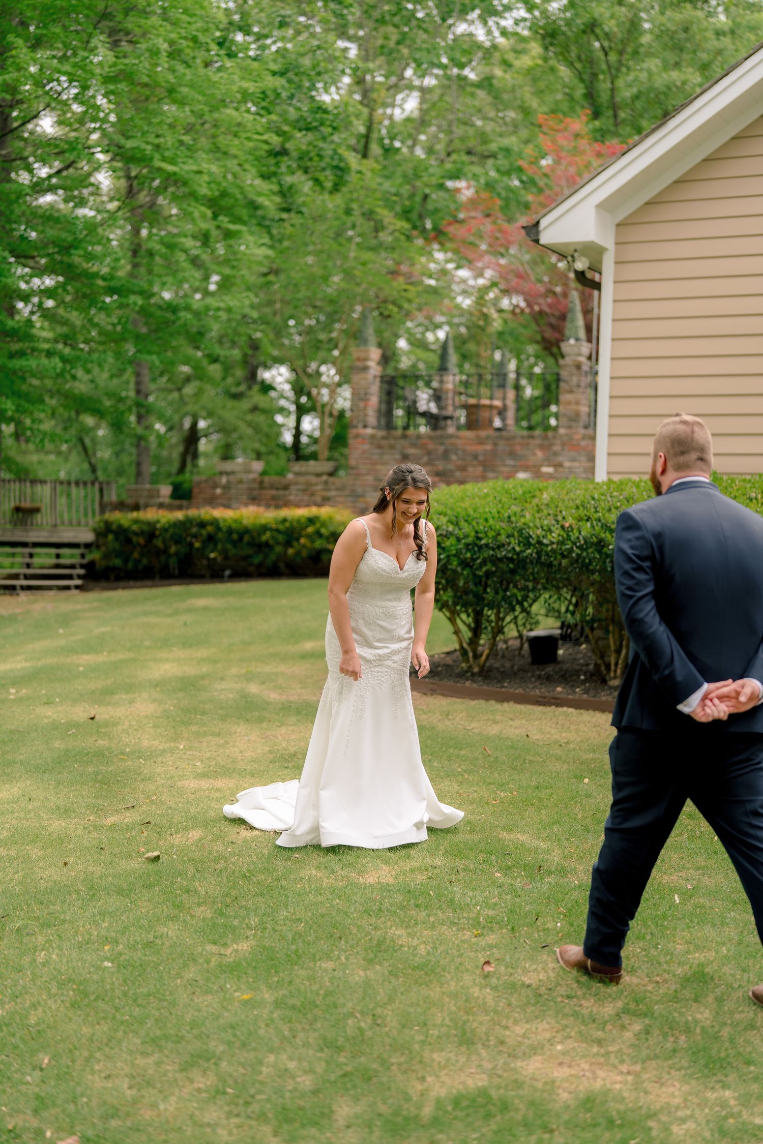 Bride in white gown smiling as groom in navy suit approaches for first look on green lawn.