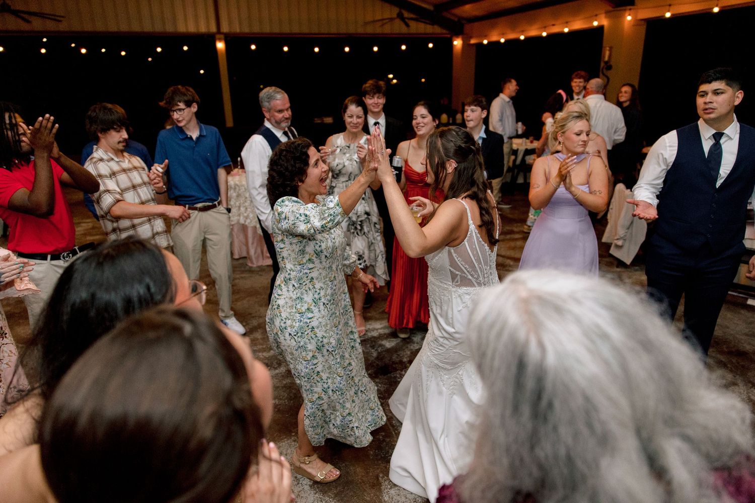 Guests surround bride dancing at festive outdoor wedding reception under string lights at night.