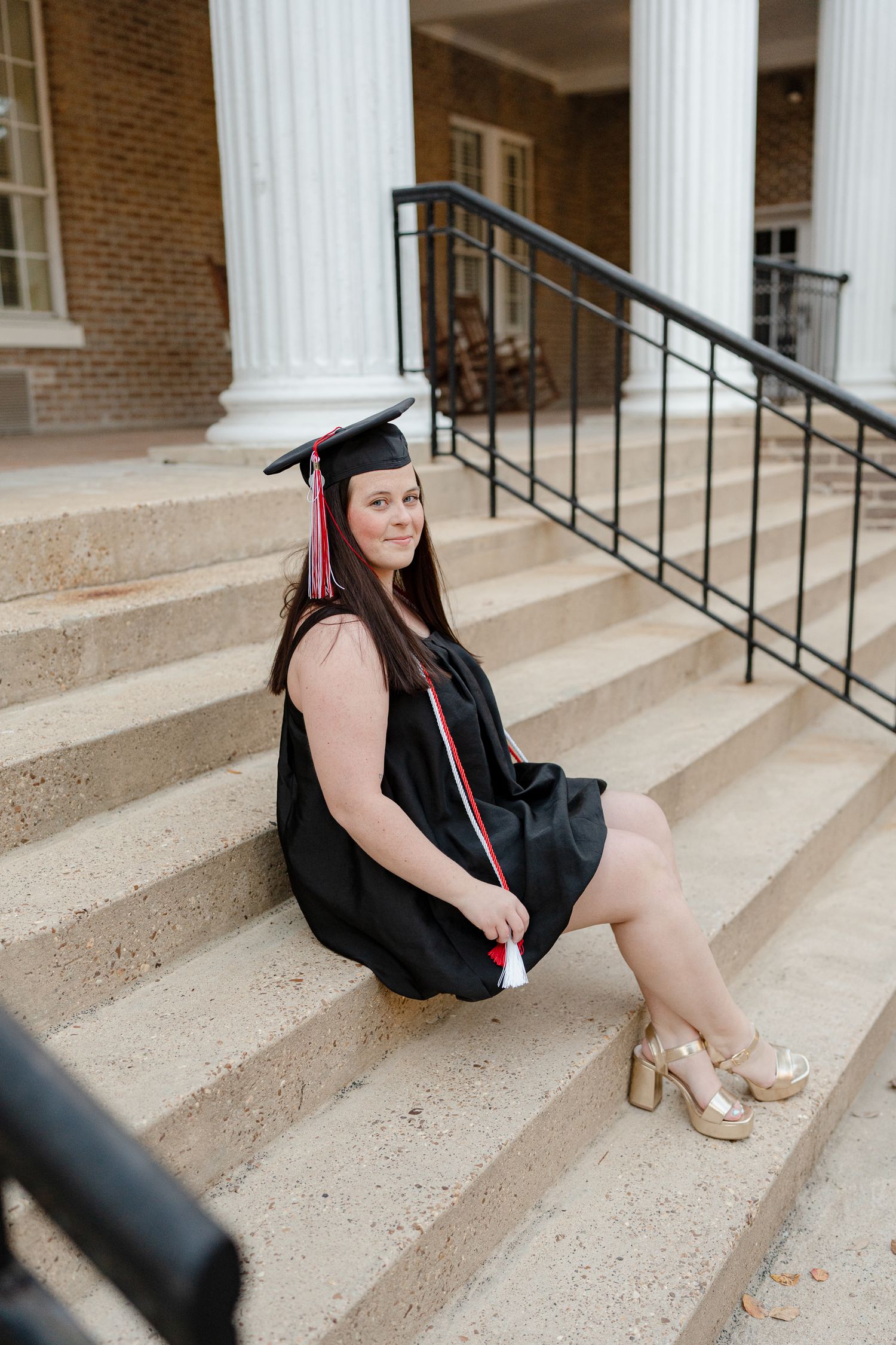 Graduate in cap and gown sits on concrete steps of brick building with white columns for graduation photos.