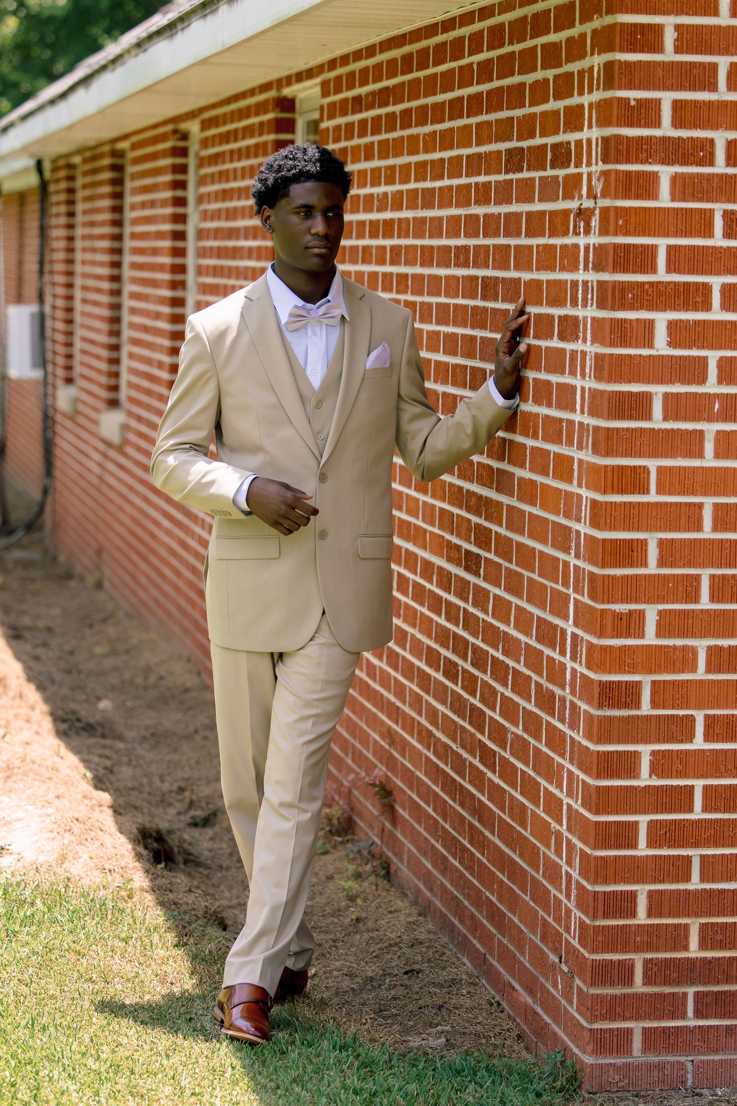Groom in a tan suit with pink bow tie poses against a red brick wall outdoors.