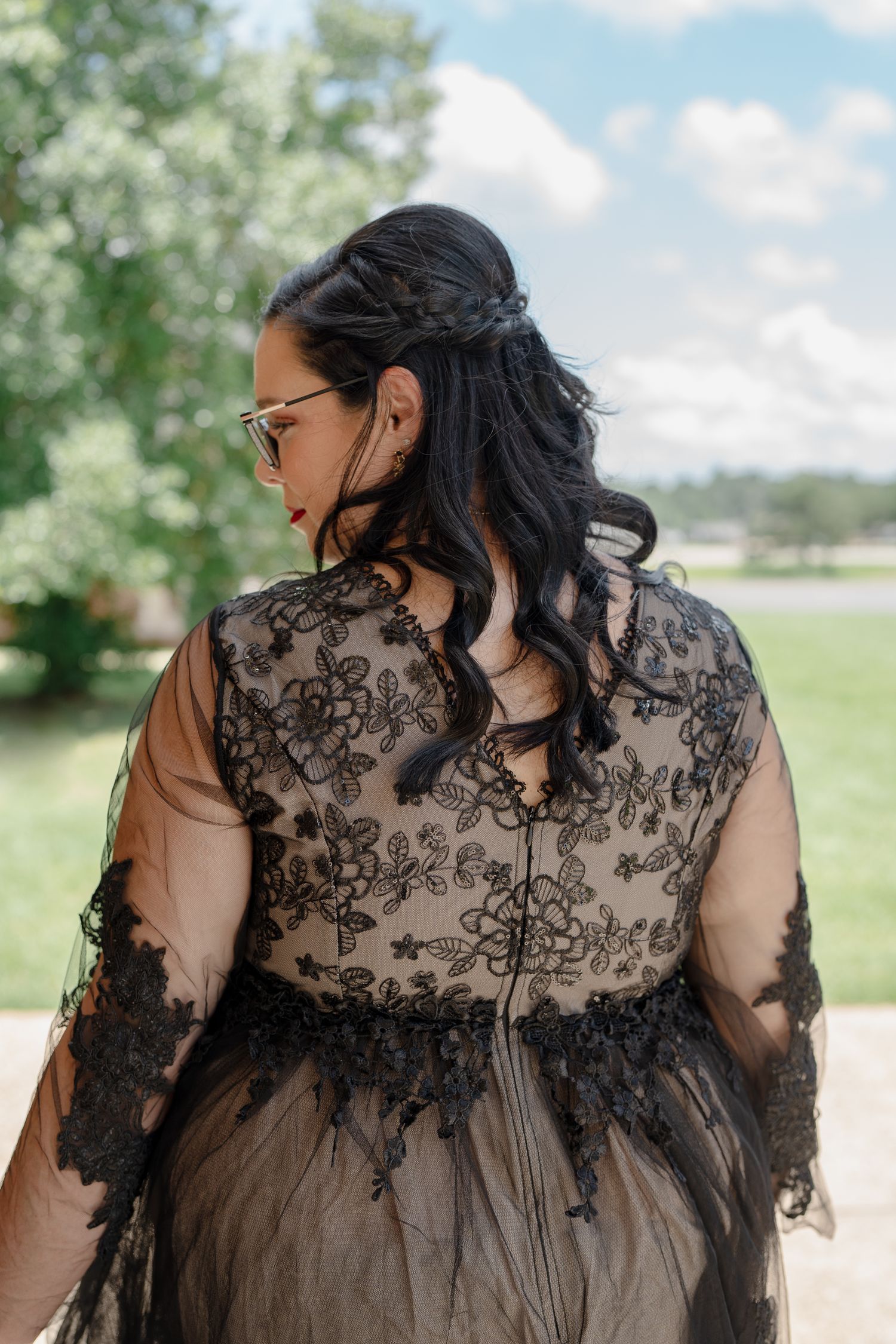 Back view of an ornate black lace dress with sheer sleeves against a sunny outdoor backdrop.