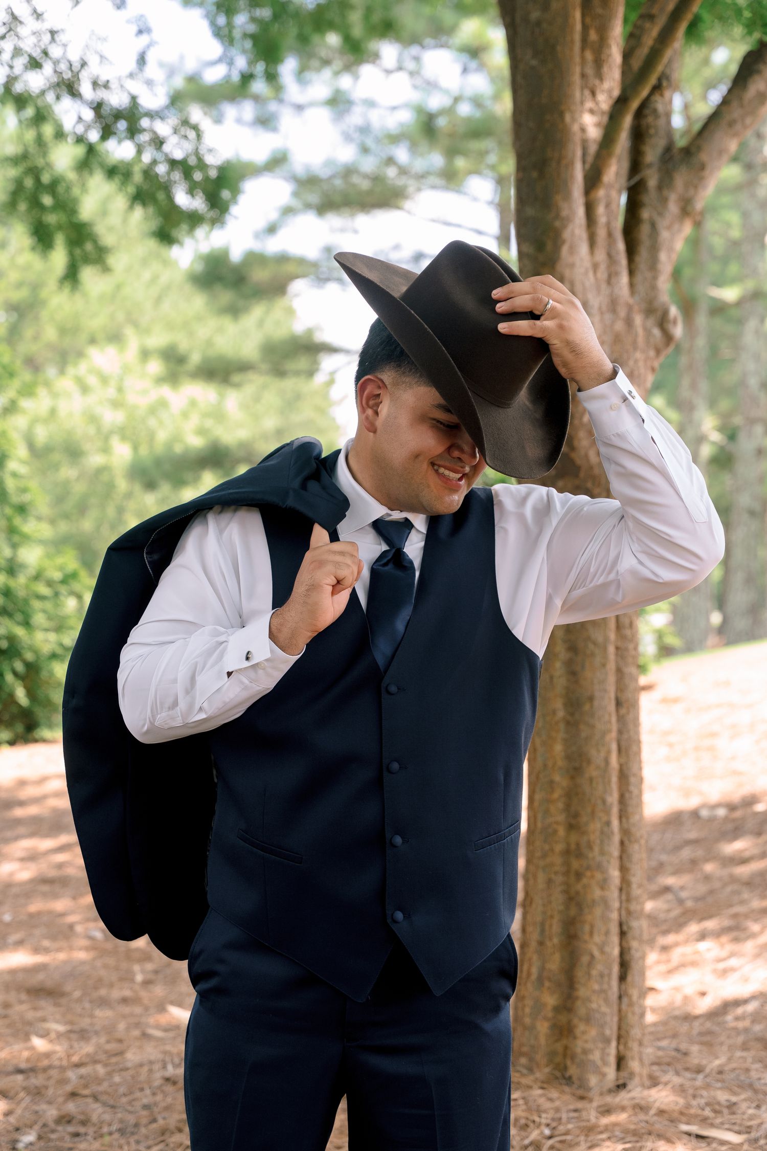 A western-style groom in a navy vest and cowboy hat poses casually in a wooded outdoor setting.