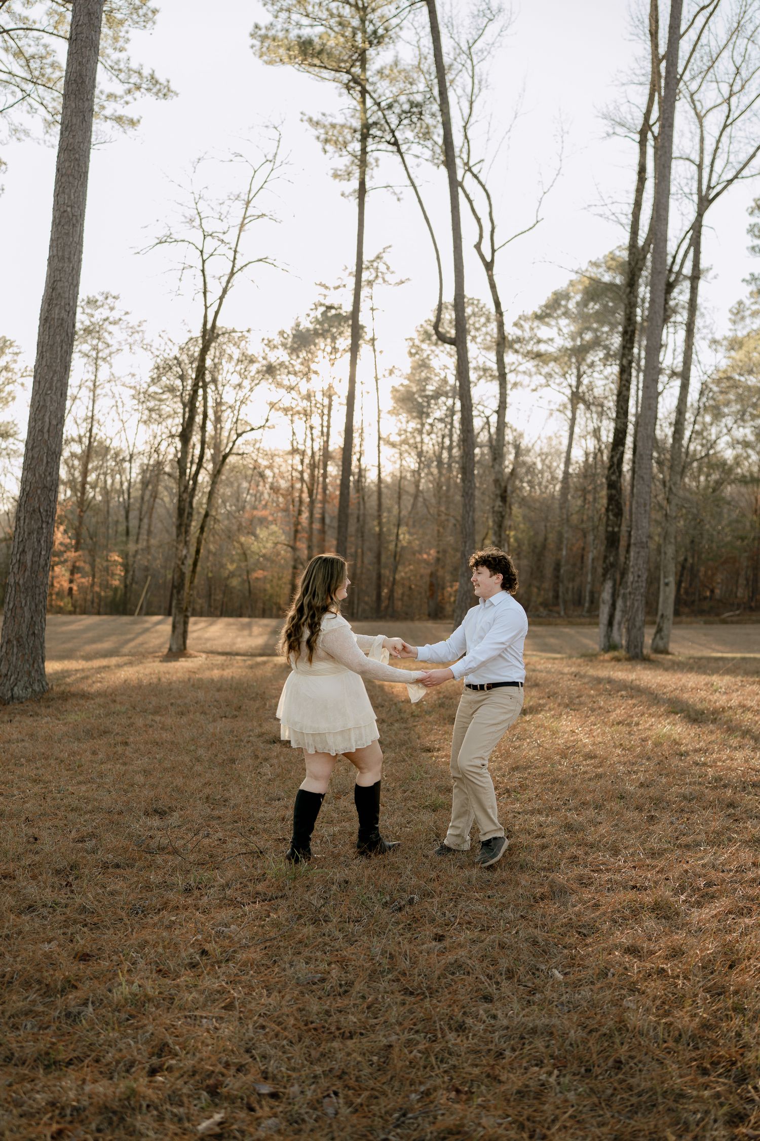 Couple holding hands and spinning joyfully in a sunlit forest clearing during golden hour.