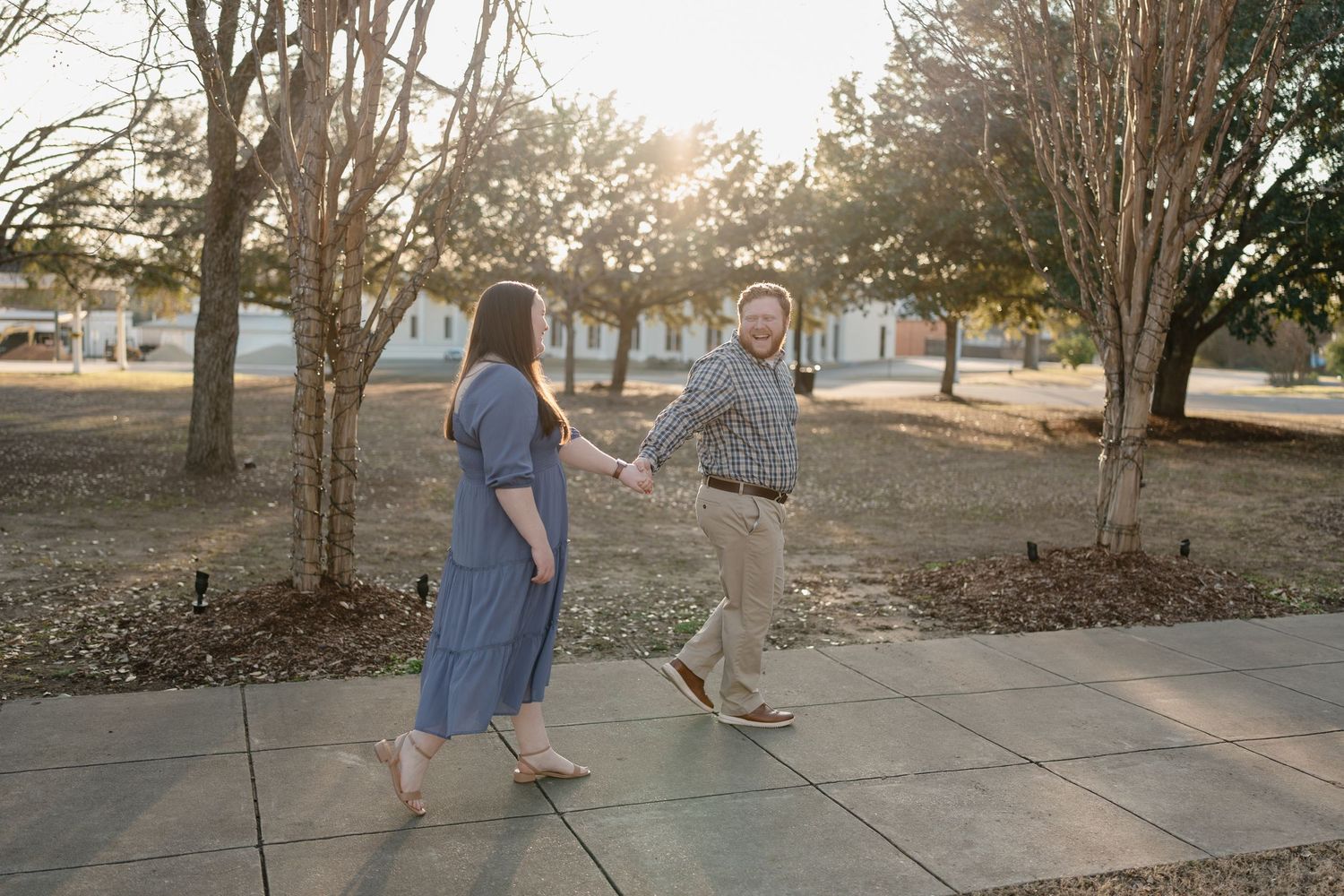 Couple holding hands walking on sidewalk among trees in warm golden sunlight outdoors.