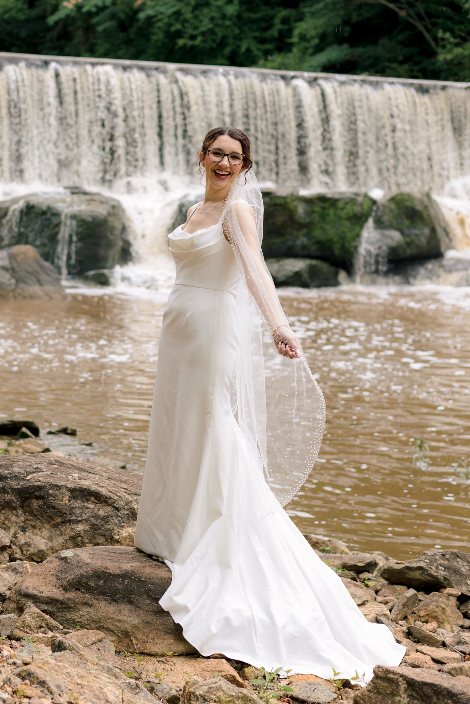 Smiling bride in elegant white gown standing on rocks in front of a cascading waterfall.