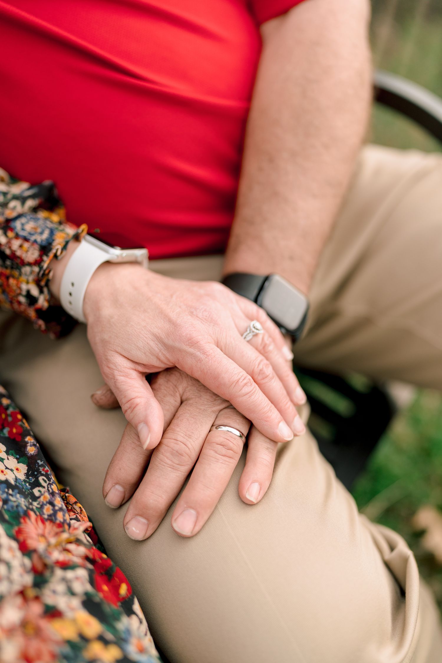 Close-up of a couple's hands resting together showing wedding rings and fitness trackers on their wrists.