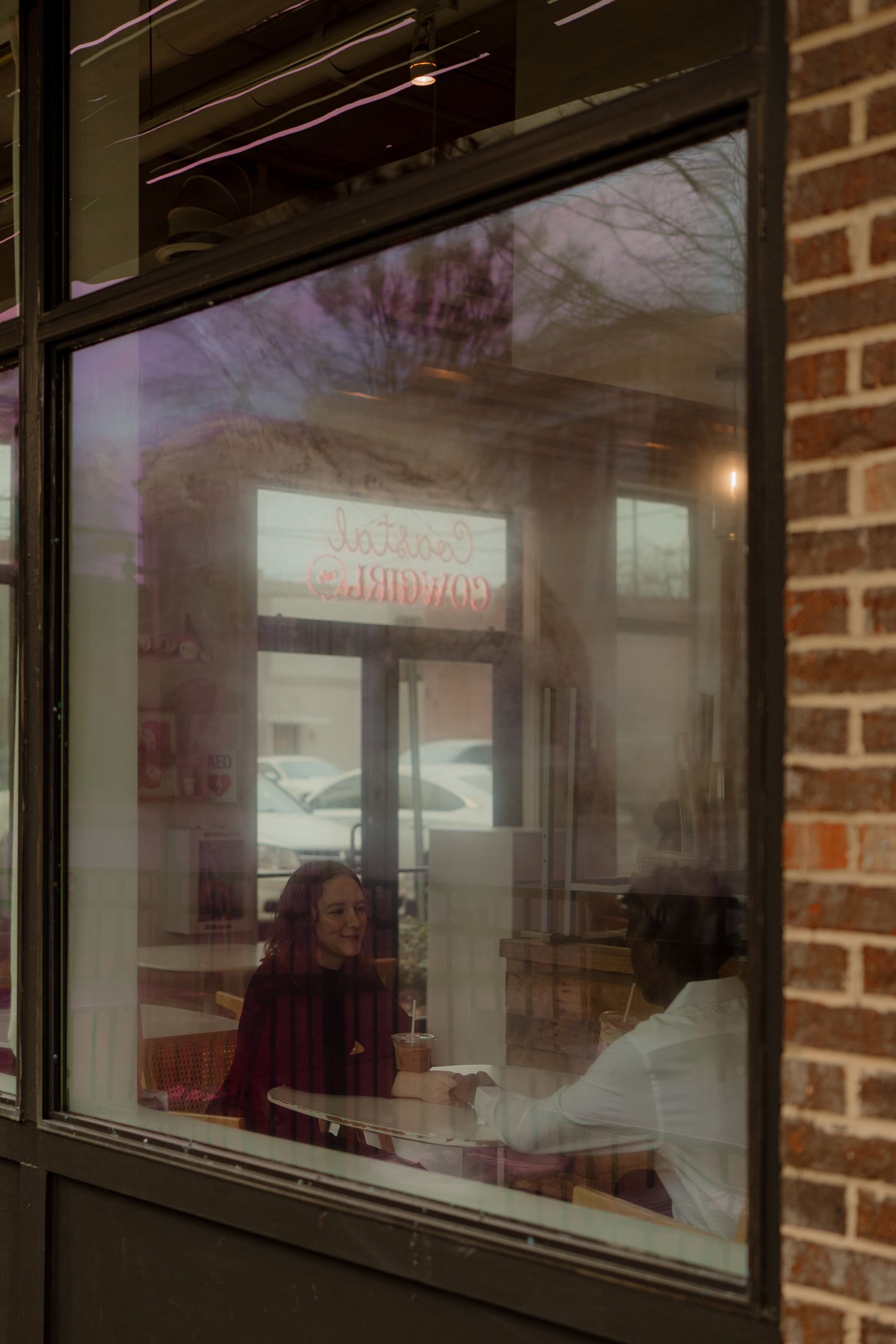 Couple sitting together inside a cozy cafe viewed through a brick-framed window at dusk.